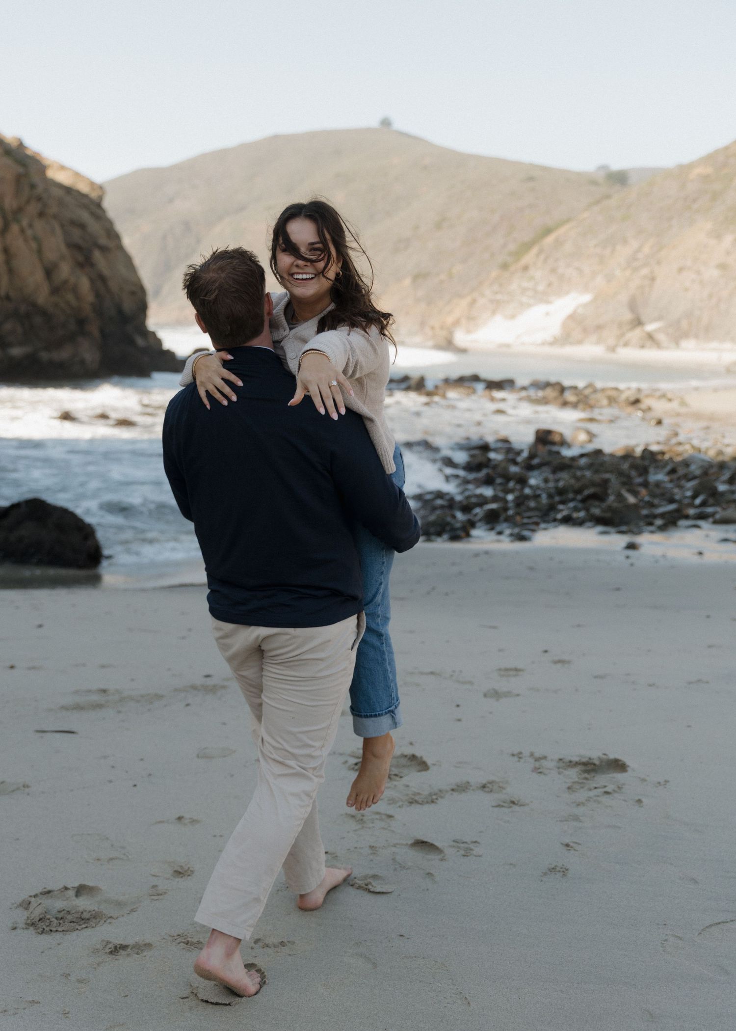 A newly engaged couple with the man holding a woman showing off her engagement ring as they walk down the beach in Big Sur, California.