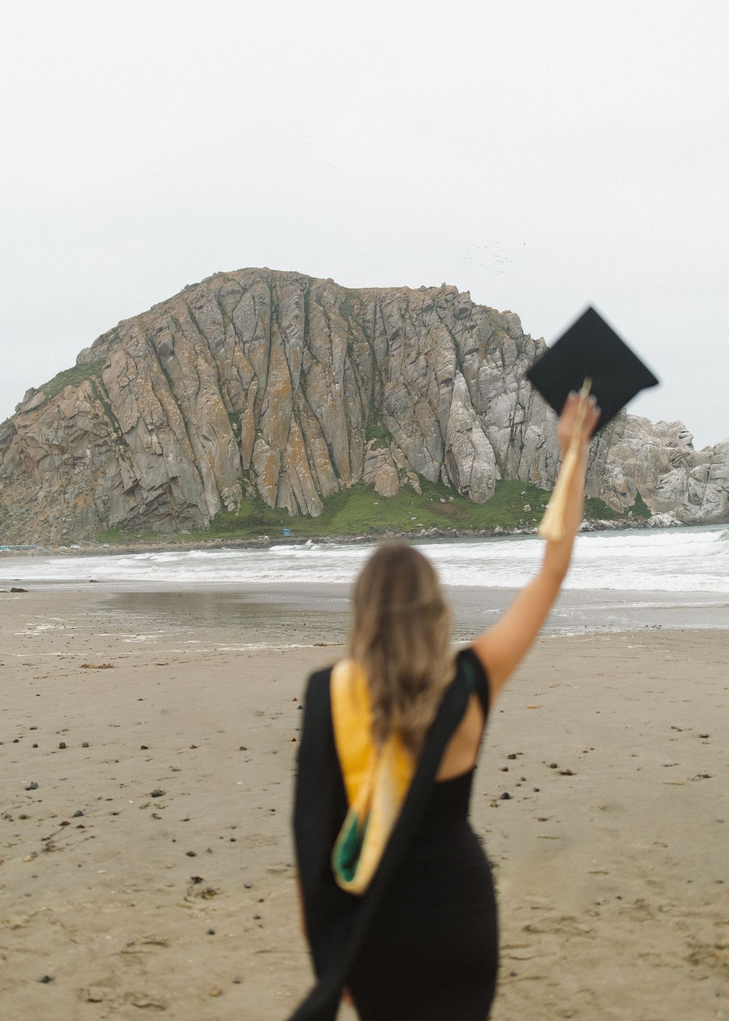 A Cal Poly San Luis Obispo graduate holds their graduation cap into the air on a California beach with Morro Bay rock.