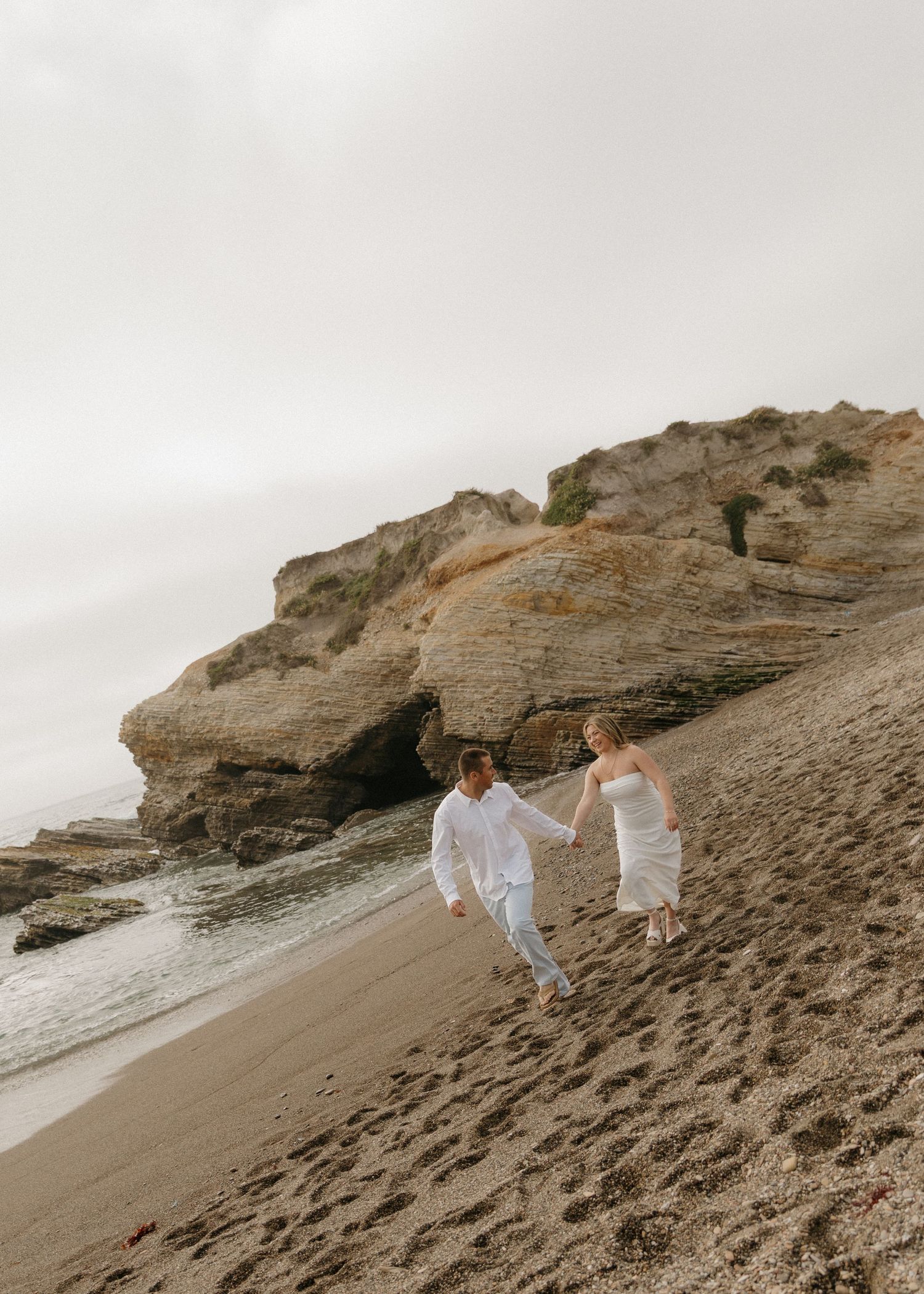 An engaged couple walks along the scenic Montana De Oro Beach in Los Osos, California with rock formations and cliffs in the background.