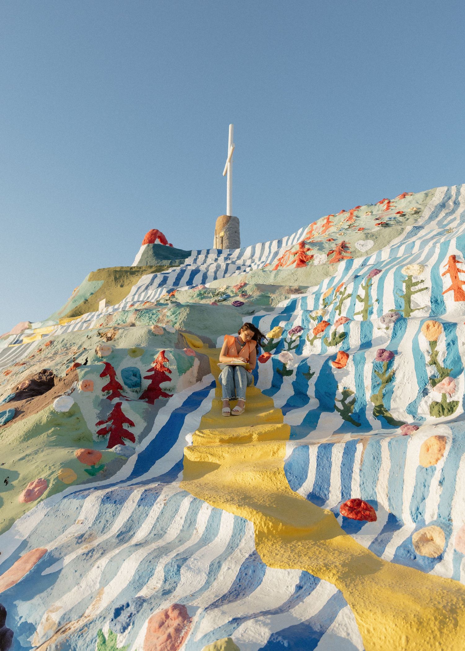 A wide shot of Salvation Mountain's painted slopes with a graduate studying her bible in Joshua Tree, California.