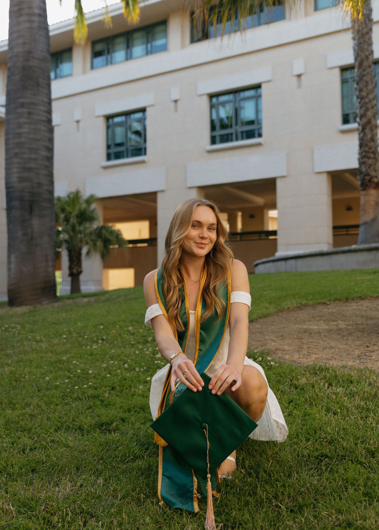 Cal Poly San Luis Obispo graduate sits gracefully holding her cap near Orfalea College of Business building with palm trees in the back.