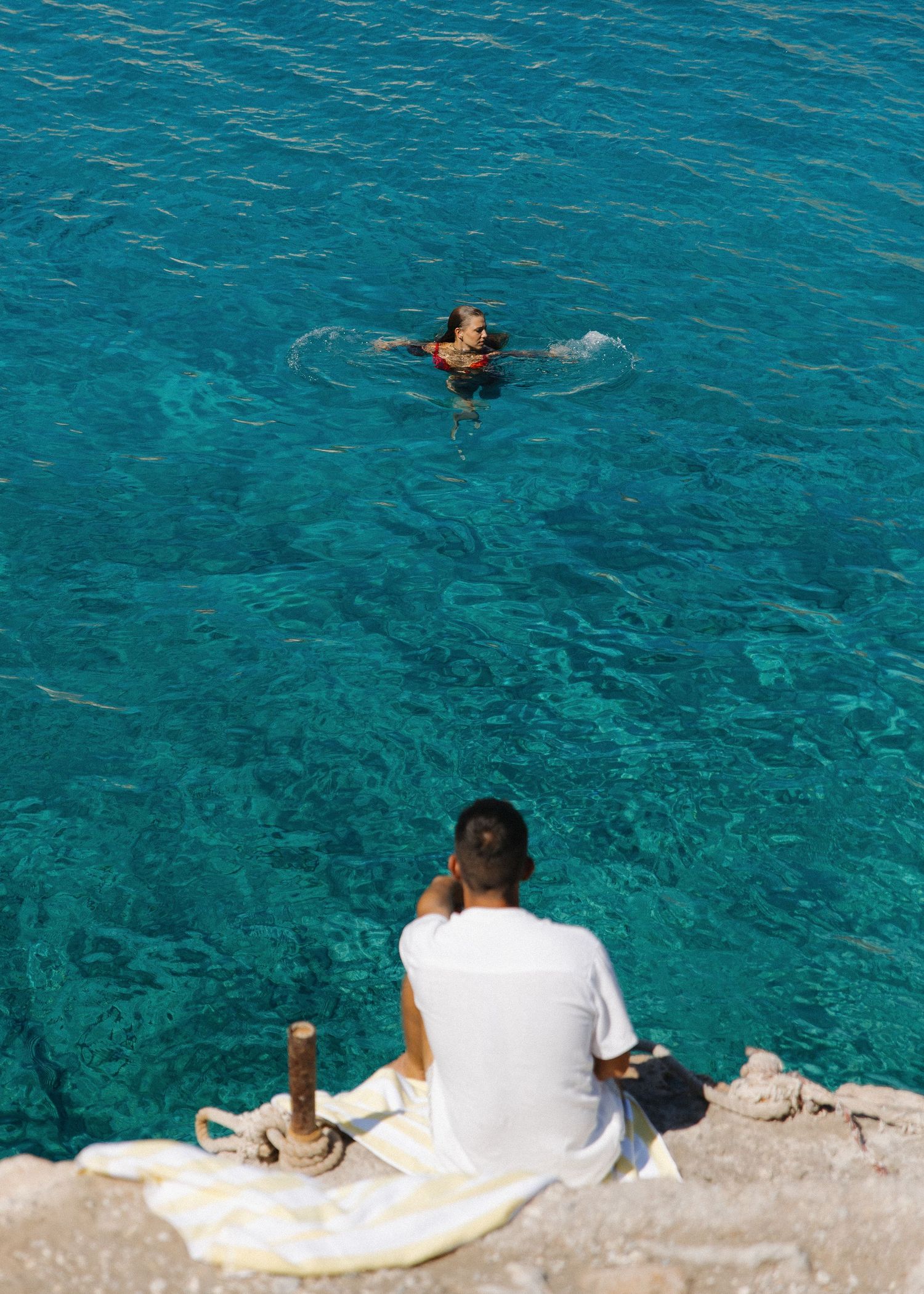 A husband relaxes on a white towel by crystal clear turquoise Mediterranean waters while he watches his wife swim in front of him.