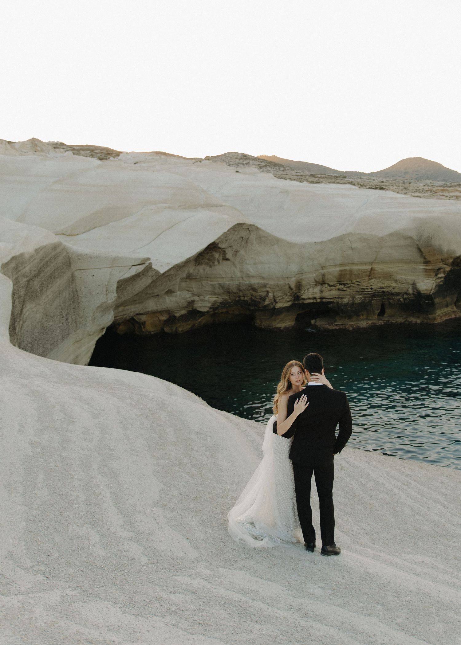 A bride and groom in wedding attire stands at the edge of Sarakiniko Beach cliffs in Milos, Greece overlooking the blue mediterranean sea.