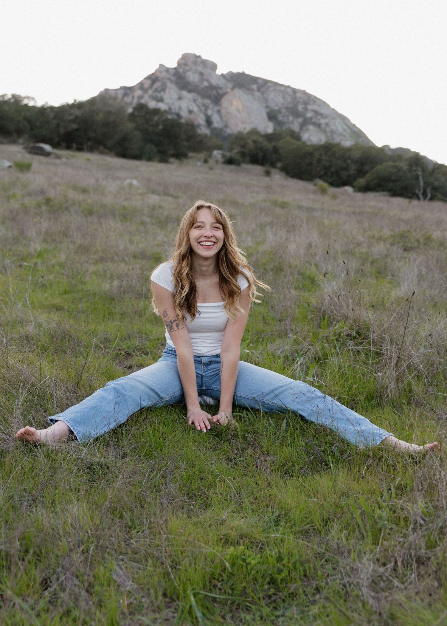 A Cal Poly San Luis Obispo graduate in casual outfit sitting playfully on grassy hillside with Bishops Peak visible in background.