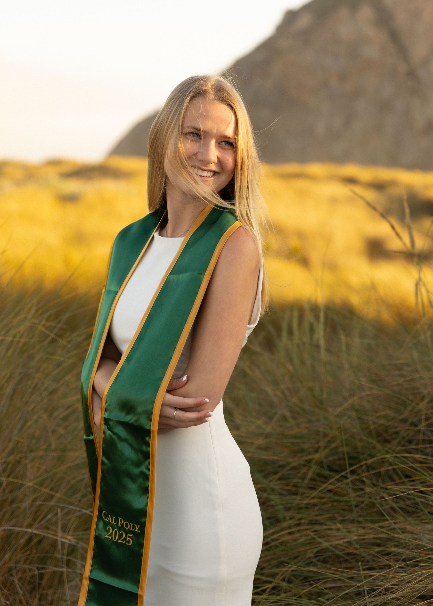 Cal Poly San Luis Obispo graduate in her stole poses in golden field in Morro Bay, California at sunset with the rock in background.