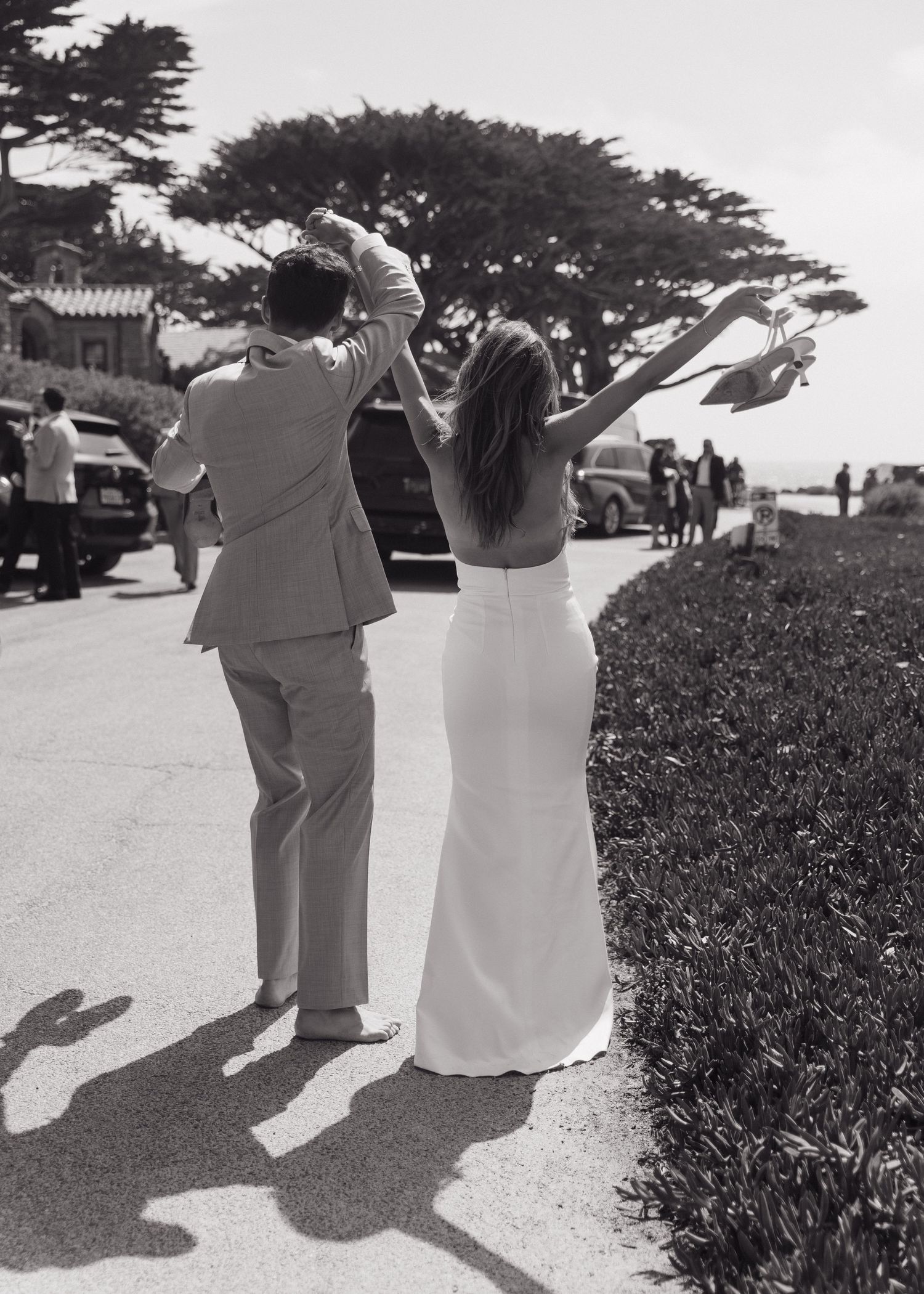 A bride and groom with their arms raised in celebration after their ceremony on a scenic coastal path in Carmel, California.