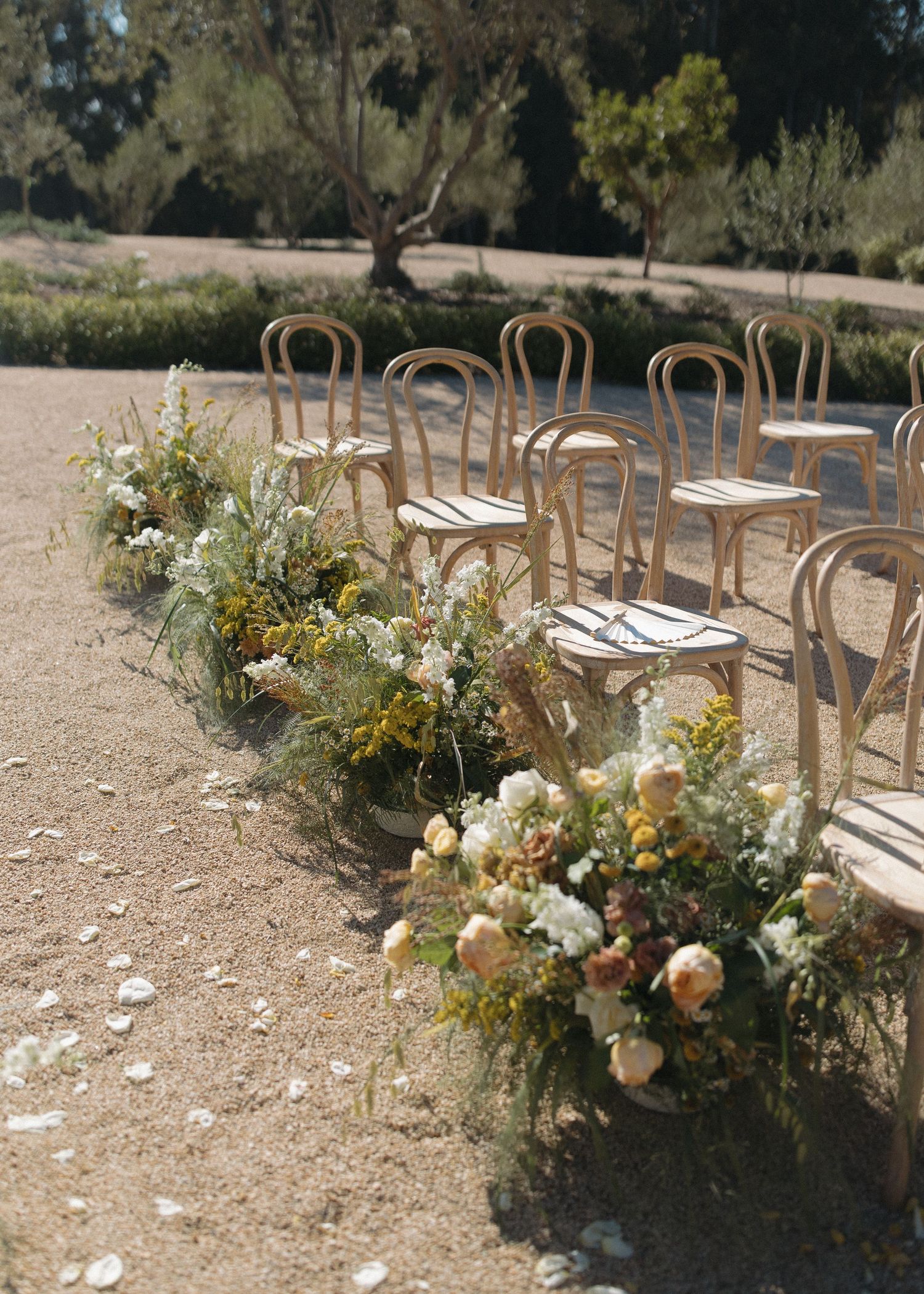 Outdoor wedding ceremony setup with wooden chairs and natural floral aisle decorations on gravel ground.