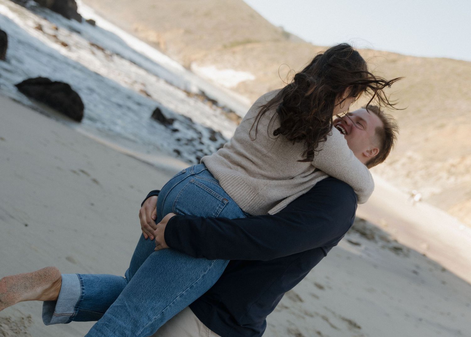 A newly engaged couple embracing while spinning on a sandy beach in Big Sur, California with waves in the background.