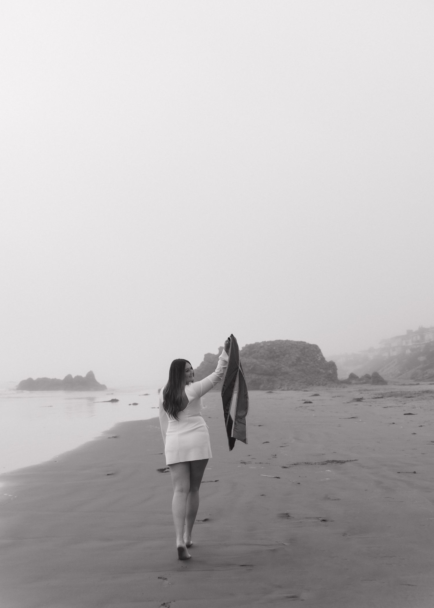 A Cal Poly San Luis Obispo graduate walks along misty Cayucos Beach, California holding up her stole in the foggy evening light.