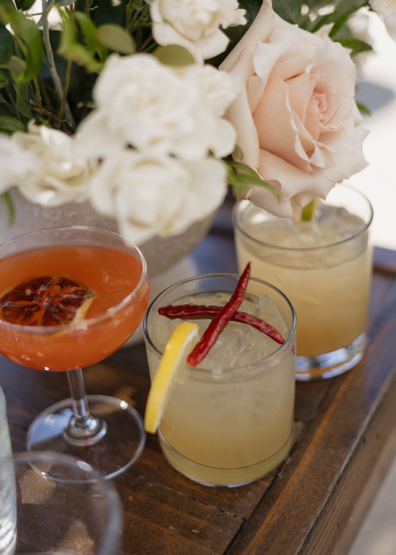 Elegant wedding cocktails with garnishes lined up on wooden surface next to white and pink roses.