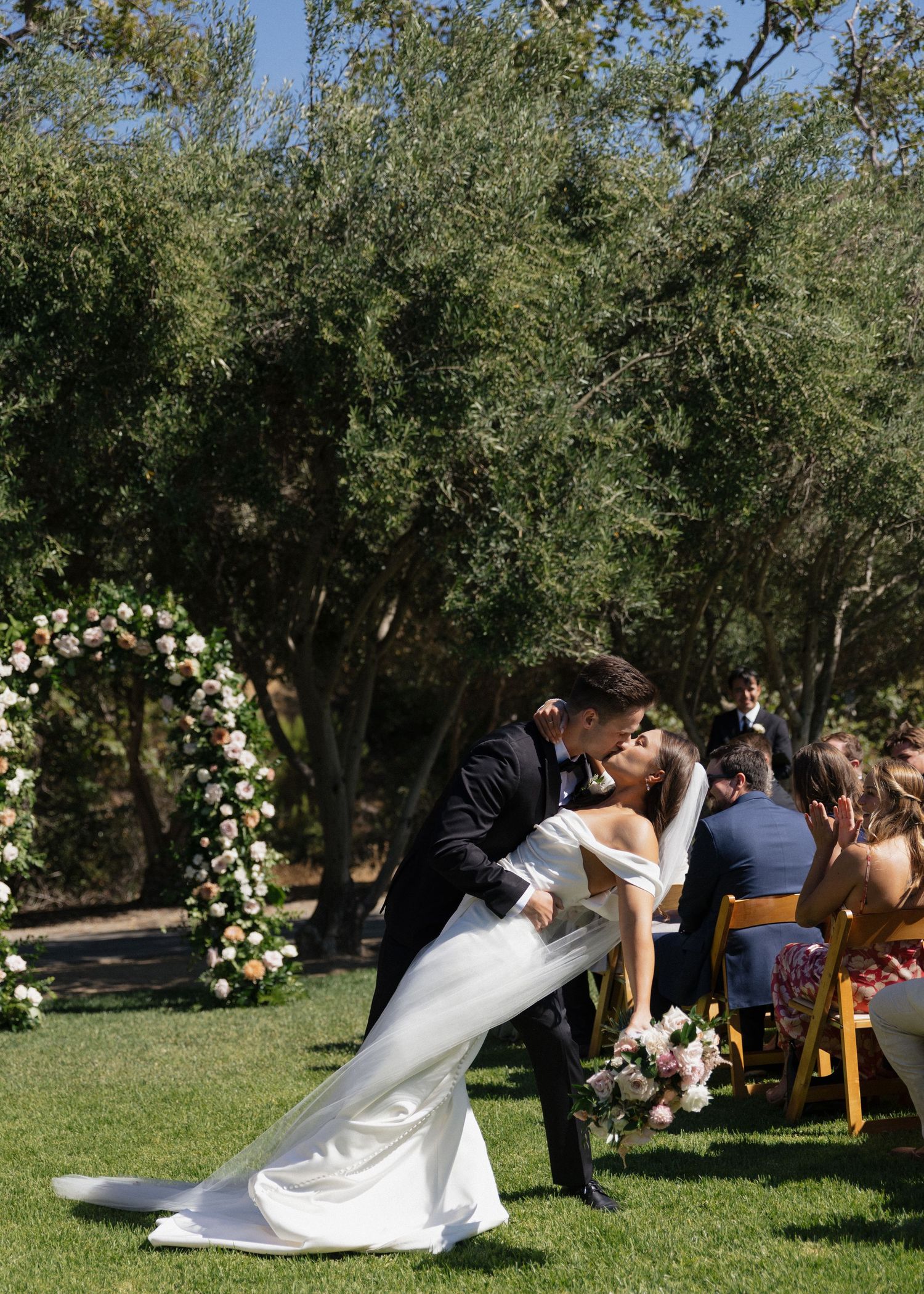 A couple sharing a dip kiss at their outdoors ceremony under a floral arch at Higuera Ranch in San Luis Obispo, California.