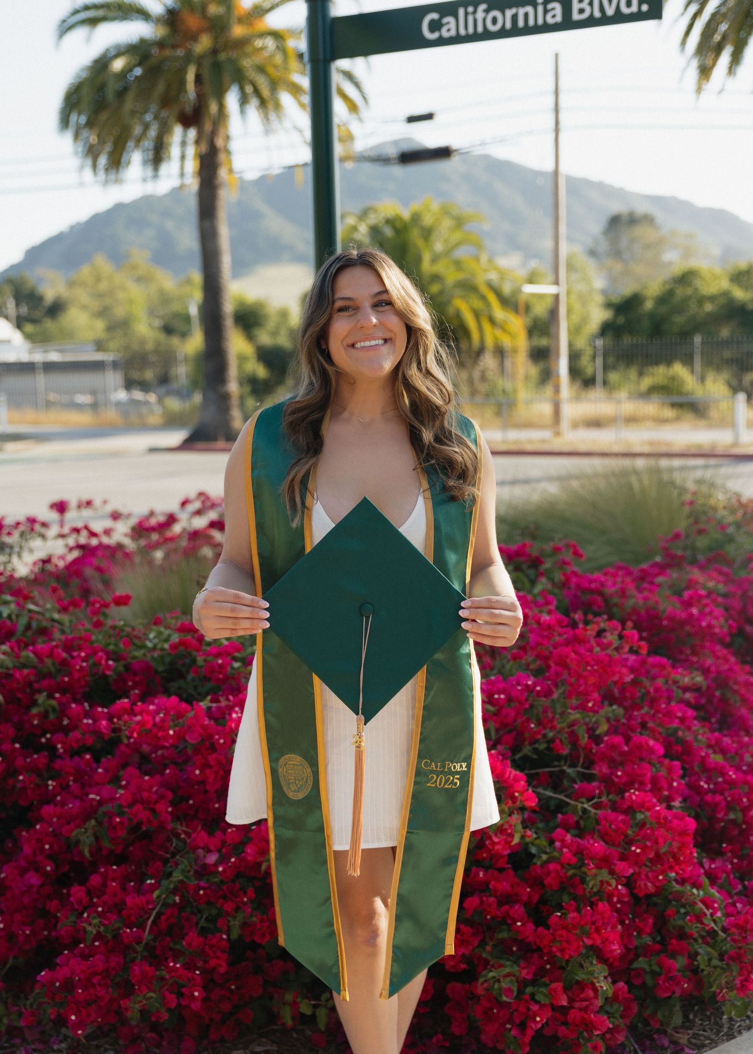 Cal Poly San Luis Obispo Graduate holding her cap poses by vibrant pink bougainvillea flowers and California Blvd street sign.