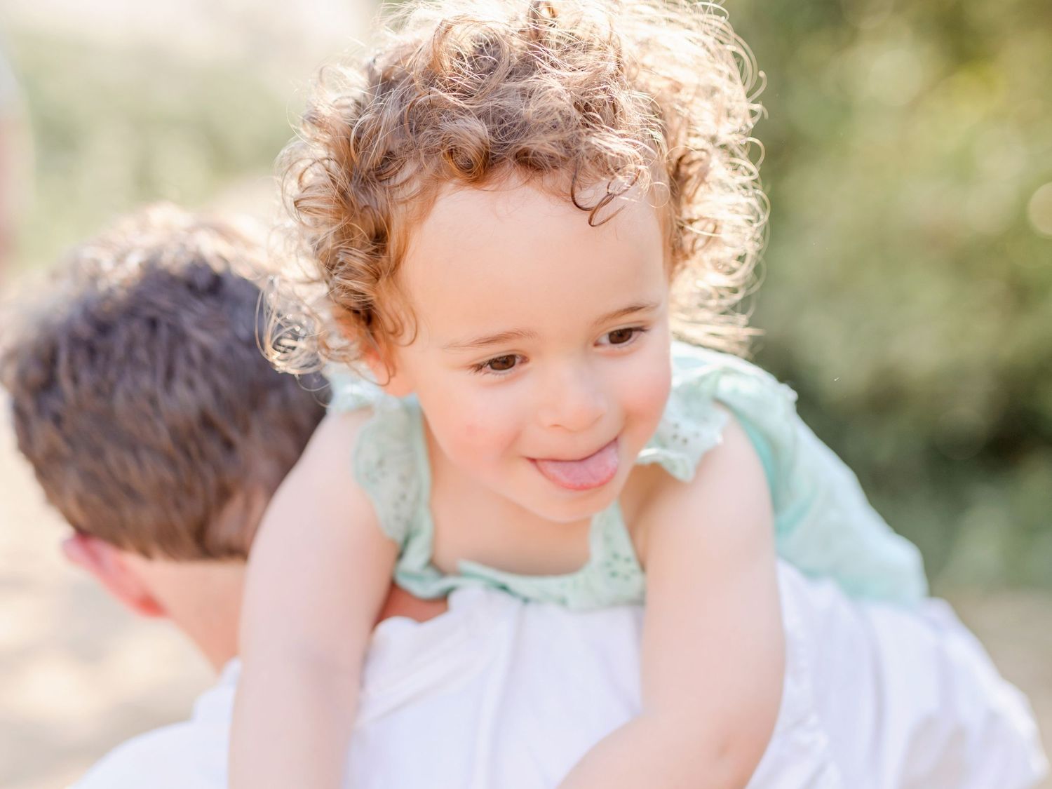 A child with curly hair gives a joyful smile while wearing a white dress shirt against a soft blurred outdoor background.