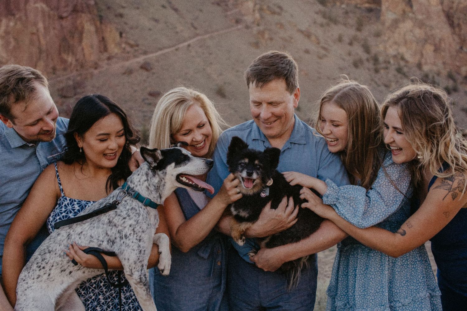 Happy family group cuddles two small dogs while standing together outdoors against a rocky canyon backdrop.