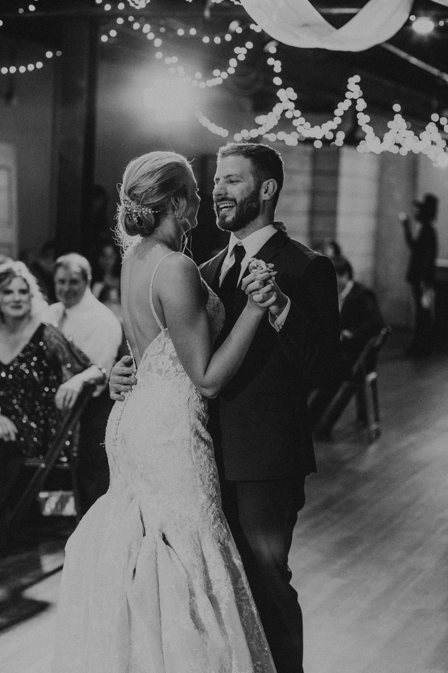 A black and white wedding photo showing a couple's first dance under romantic string lights at their reception.