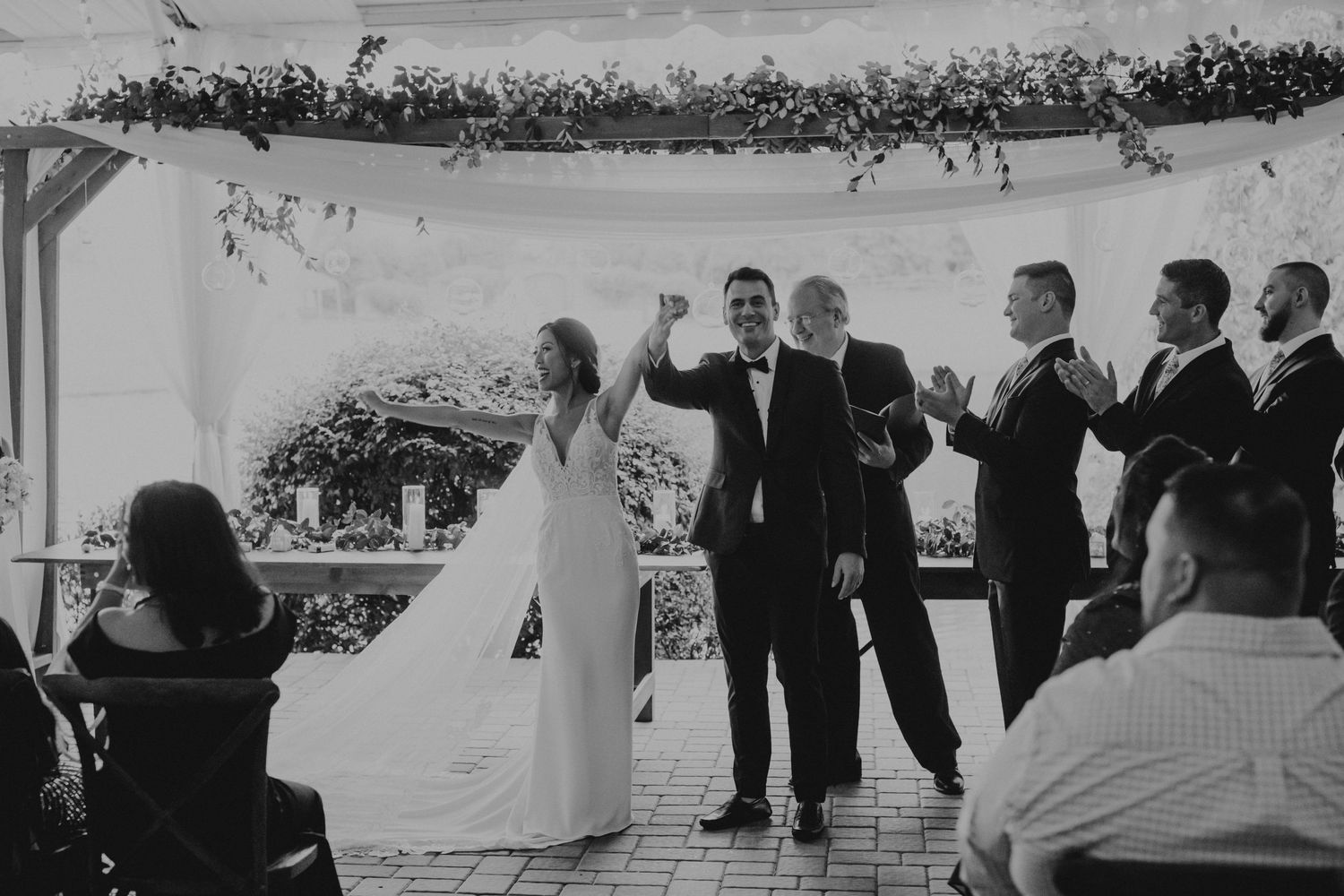 A wedding celebration taking place under a white canopy with guests gathered around on a sunny outdoor patio.