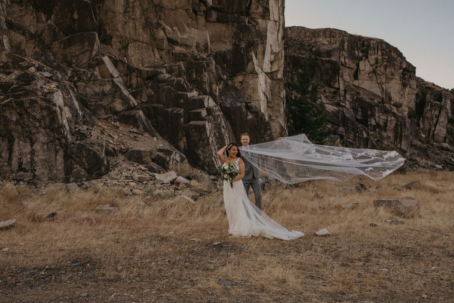 A couple in formal wedding attire embrace while a long veil flows dramatically against rocky cliff backdrop at sunset.