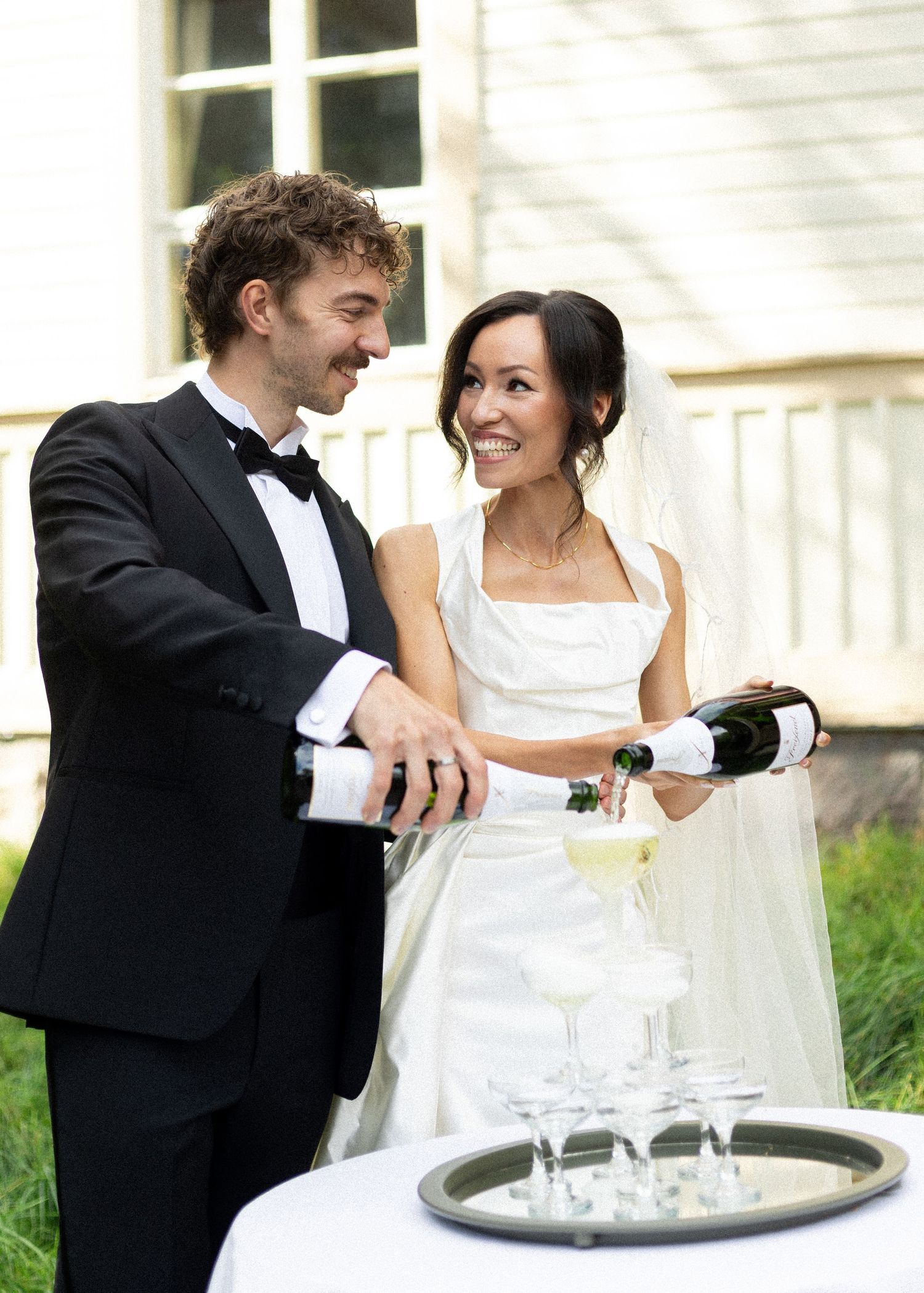 A couple in wedding attire share a joyful moment while pouring champagne into glasses on their special day.