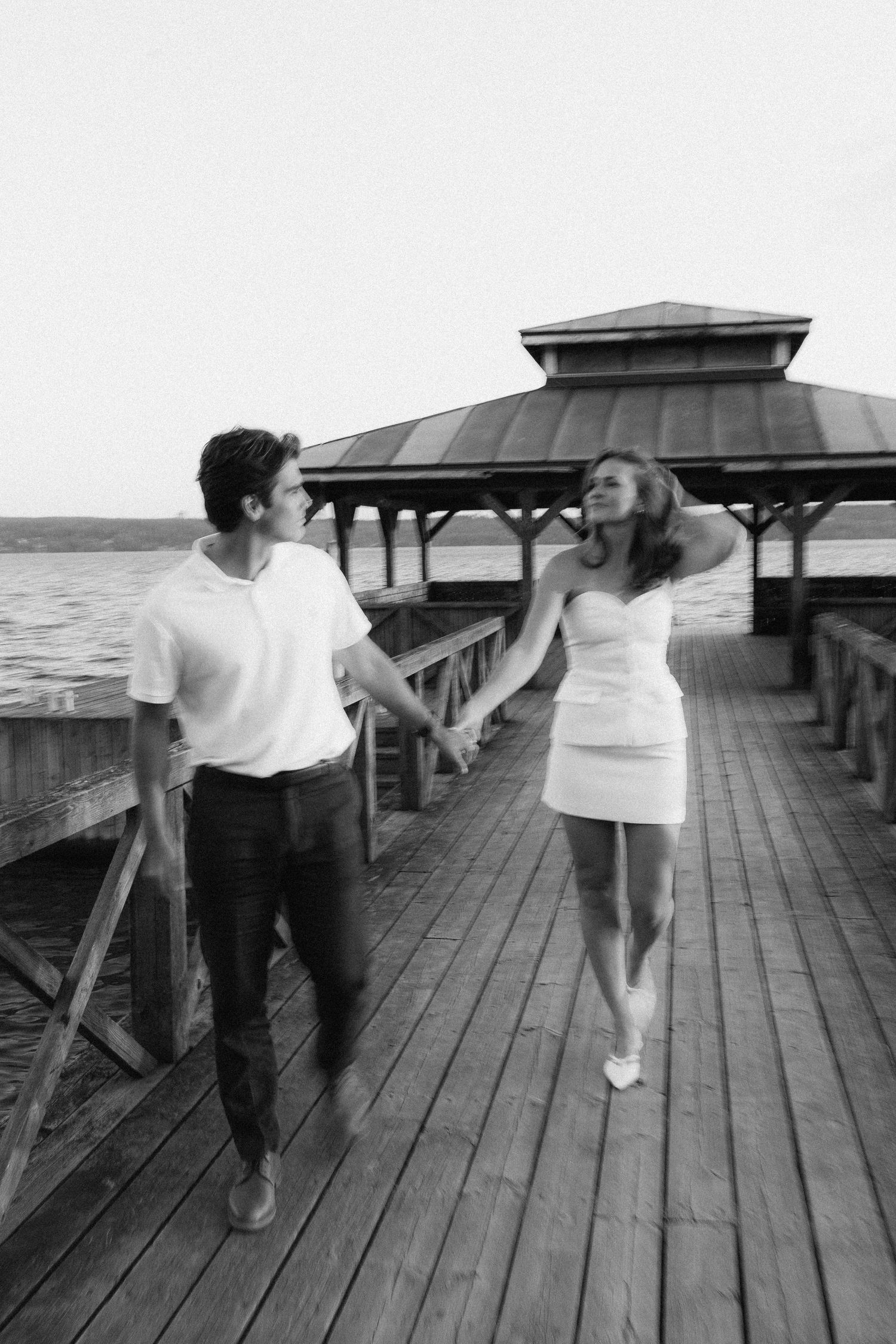 Two people in white shirts dance playfully on a wooden pier overlooking a serene waterfront.