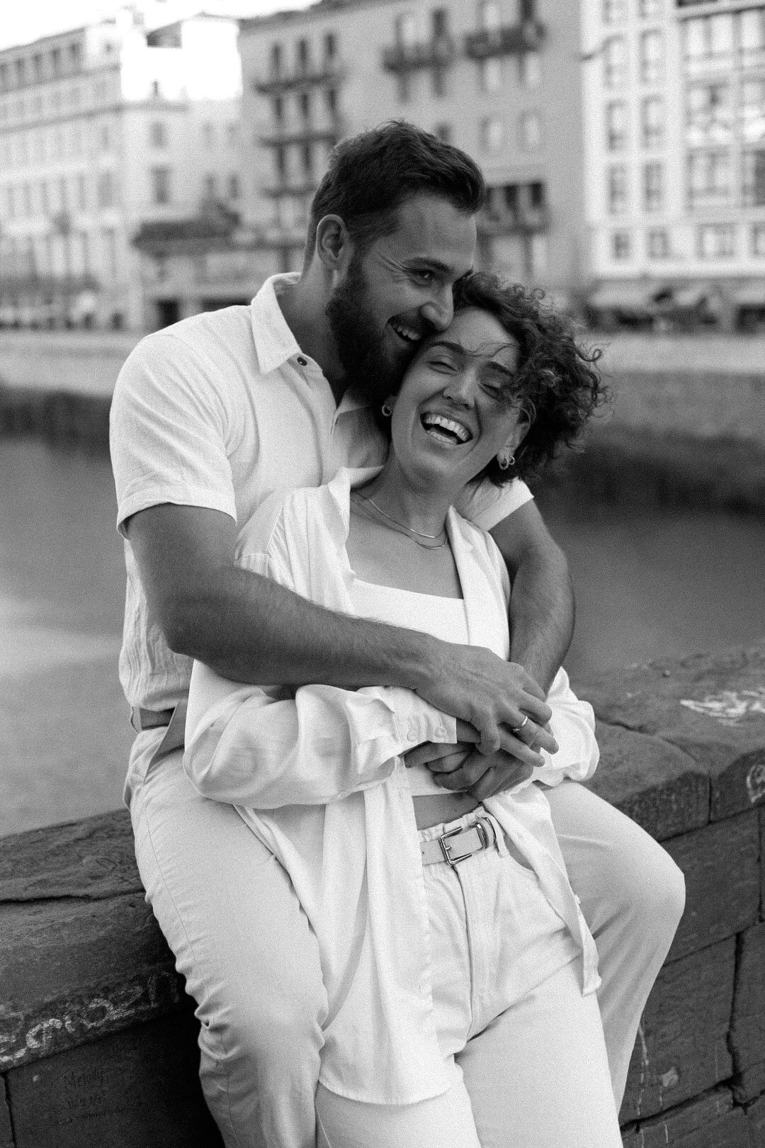 Black and white photo of a happy couple in white outfits sharing a joyful embrace while sitting by a waterfront in Florence.
