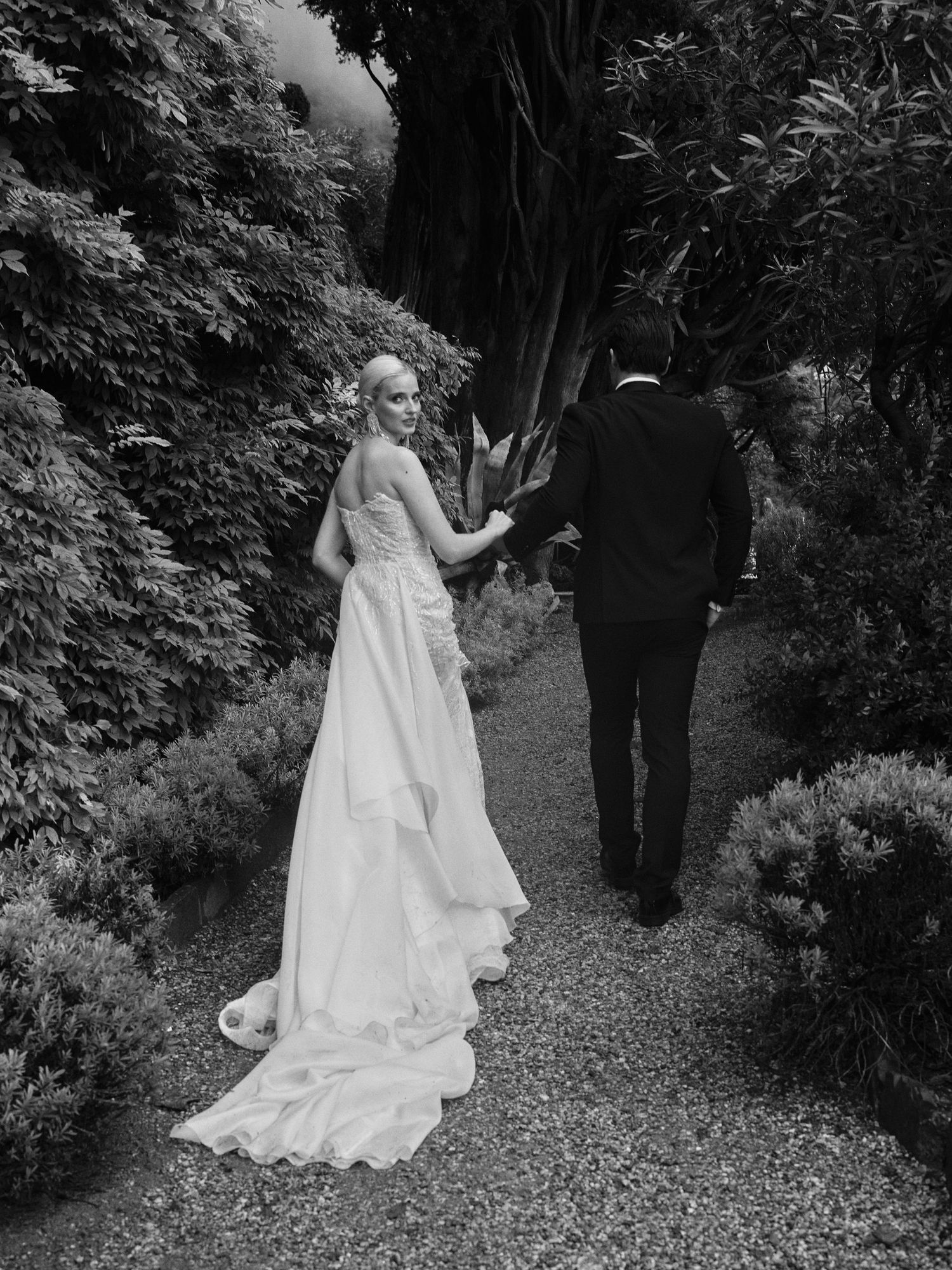 A couple in wedding attire walk along a gravel garden path surrounded by lush greenery in black and white.