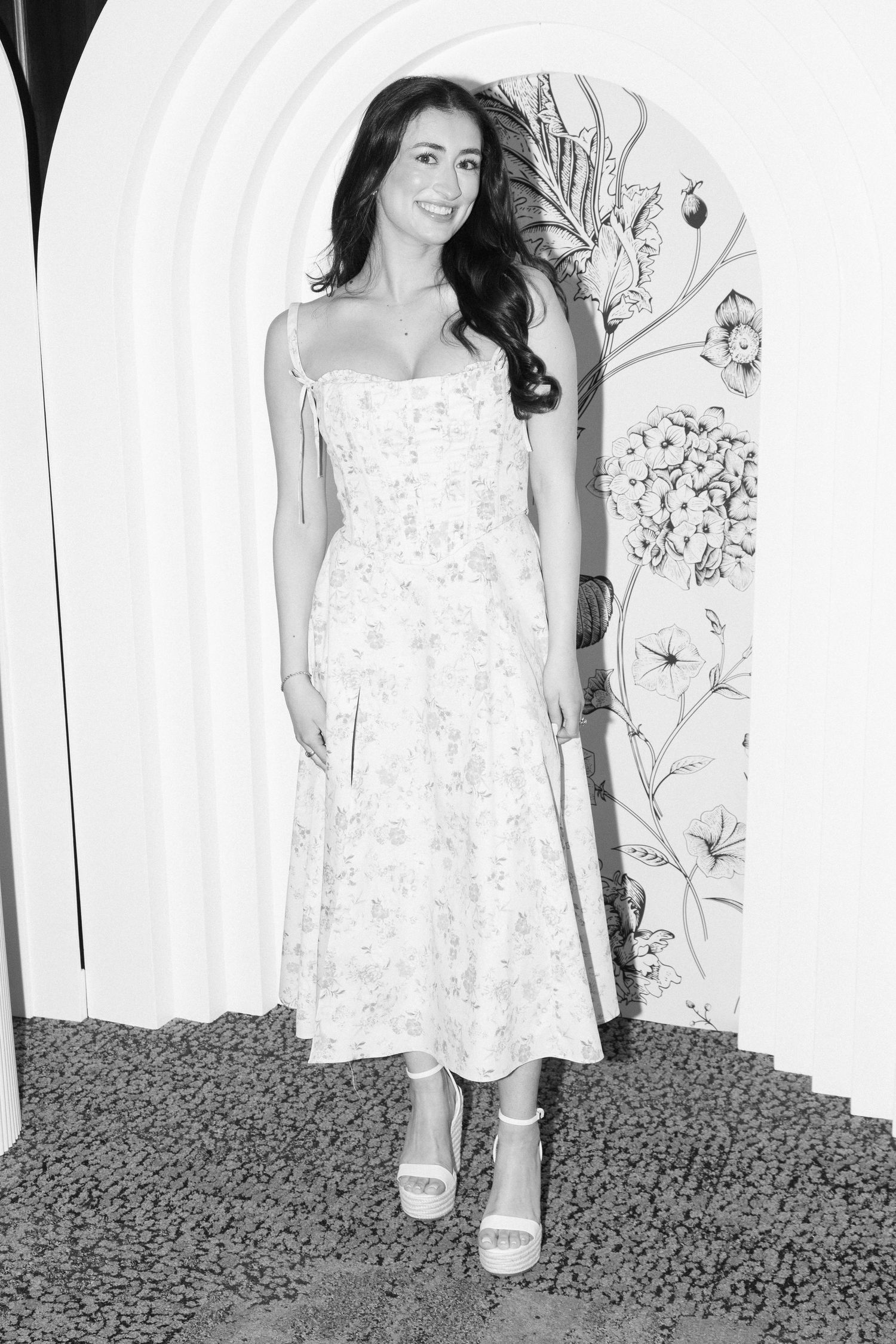 Woman in floral sundress poses against decorative wallpaper in black and white photograph.