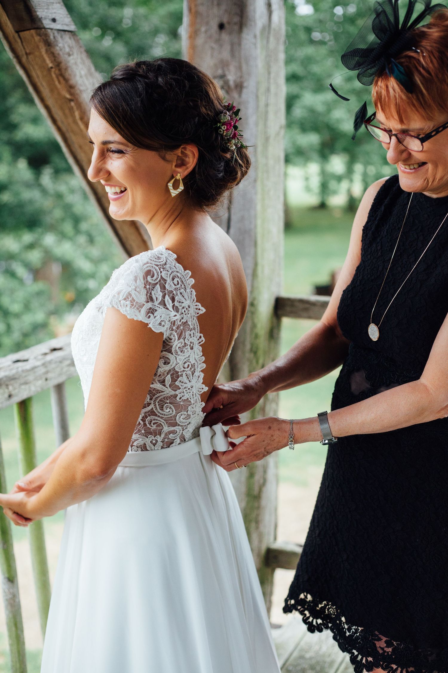 A bride in a lace back wedding dress smiles while getting ready on a rustic porch.
