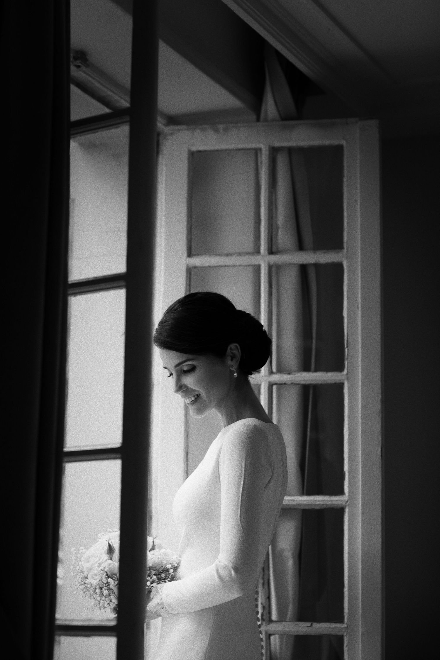 Black and white photo of a bride in silhouette standing by a window.