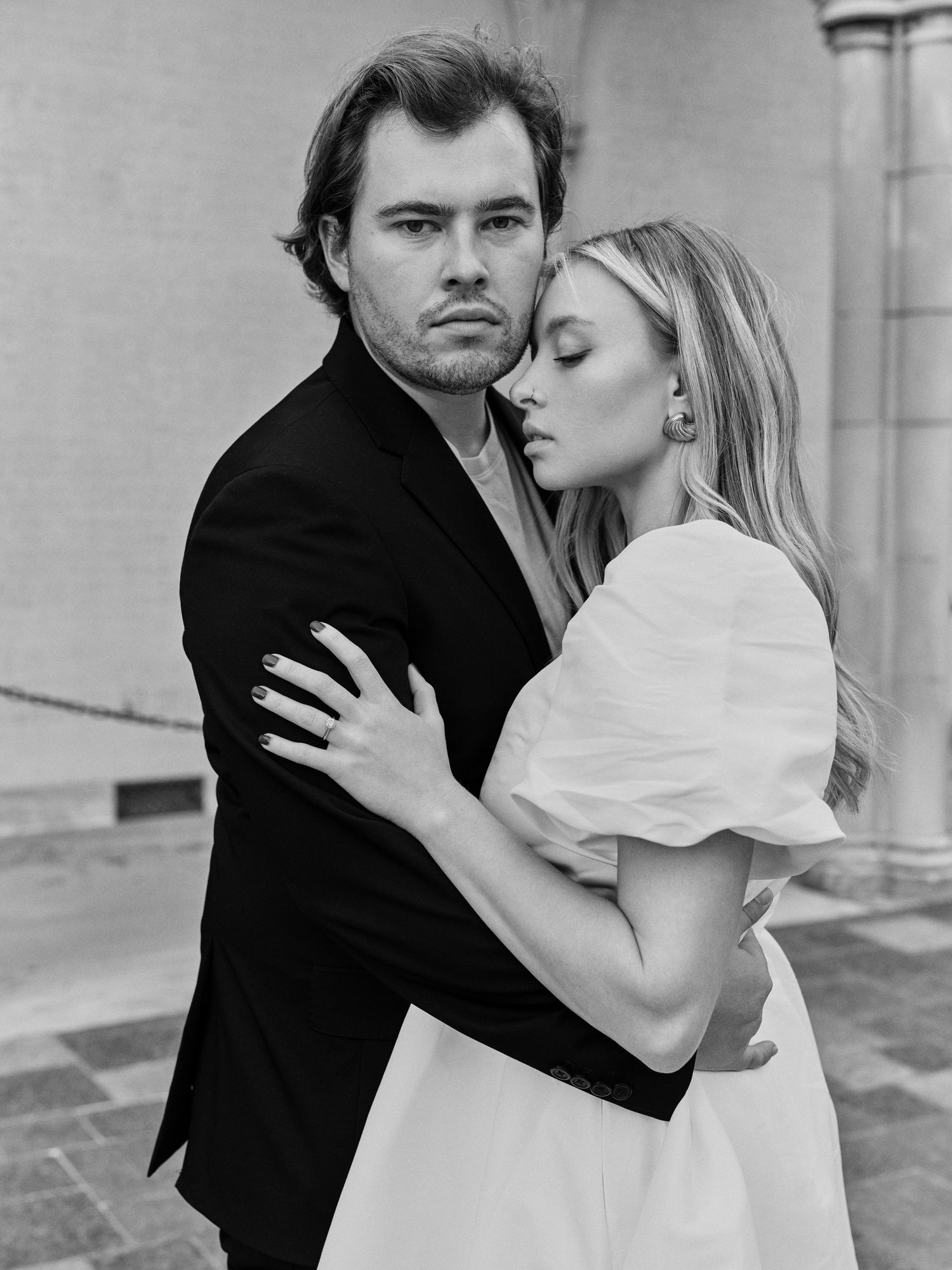 A dramatic black and white portrait of a couple in formal attire against a light wall.