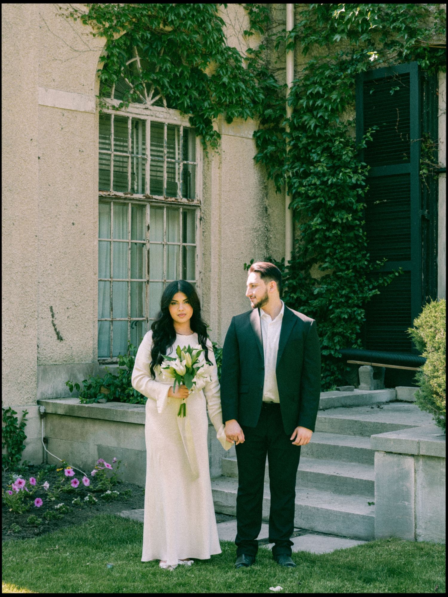 Couple in wedding attire standing on stone steps with ivy-covered walls behind them.