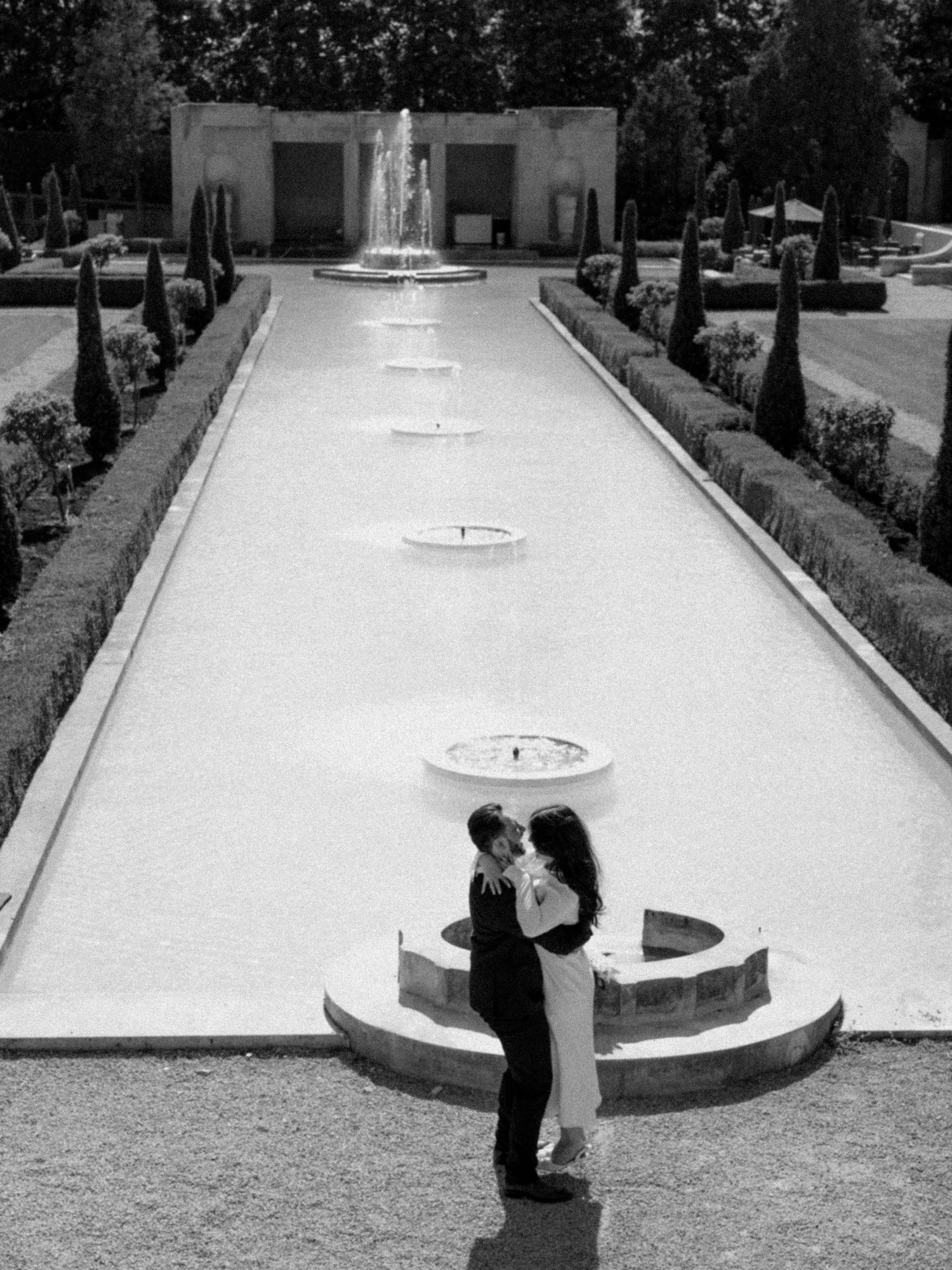 A black and white photo of a couple embracing by a formal garden fountain.