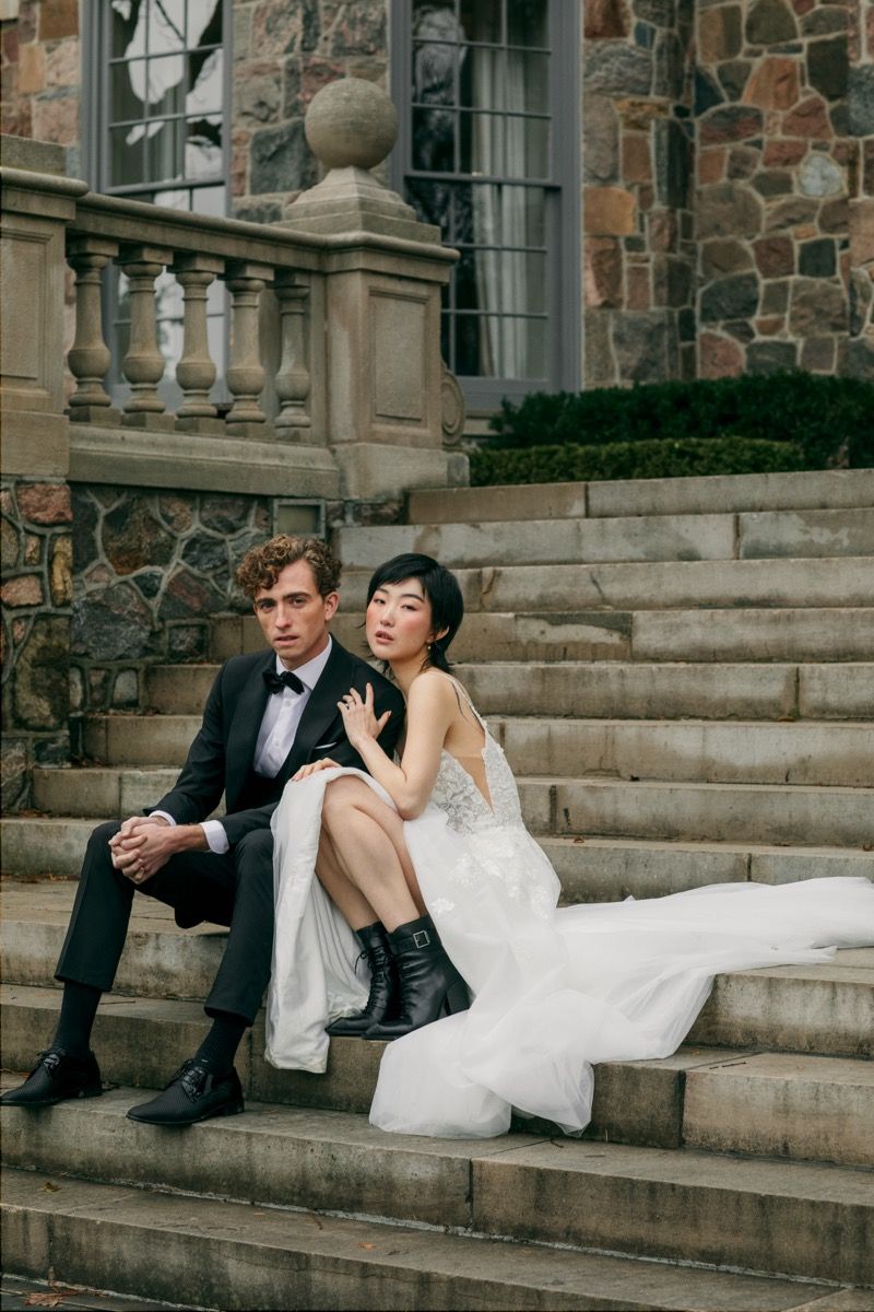 A couple in wedding attire sits casually on stone steps in front of a rustic building with black combat boots.