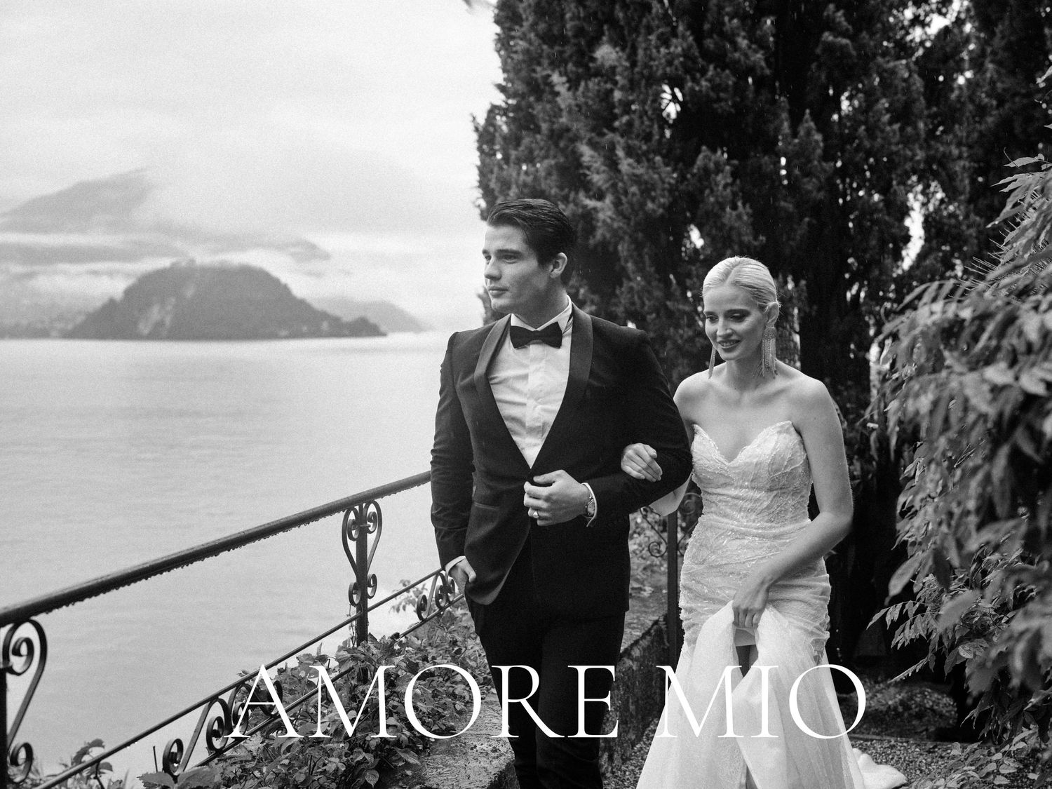 A newlywed couple stands on an elegant lakeside terrace in the Alps with mountains and cypress trees in the background.