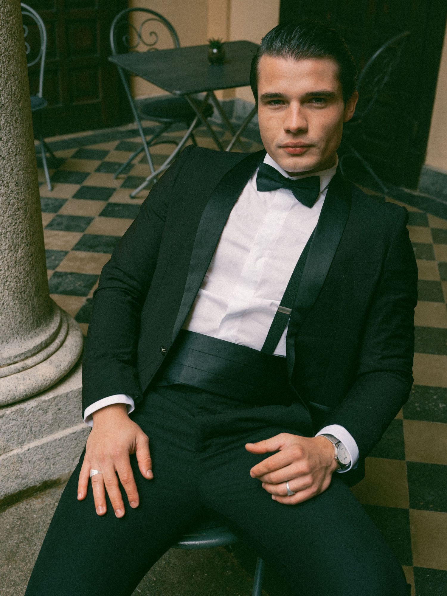 A dapper elegantly dressed person in a black tuxedo and bow tie sits confidently on a checkered floor.