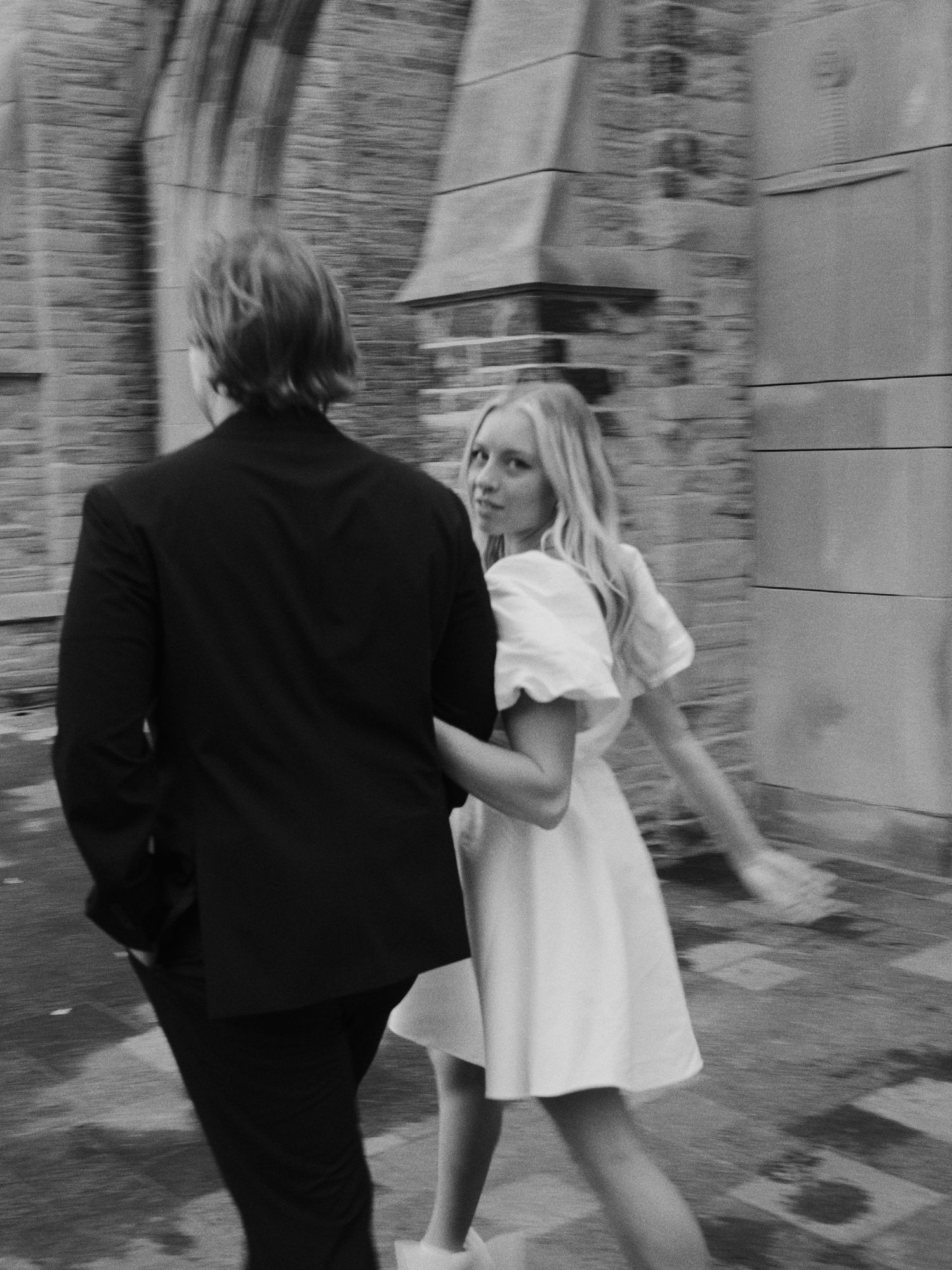 Black and white photo of couple walking near stone building, she in white dress glancing back.