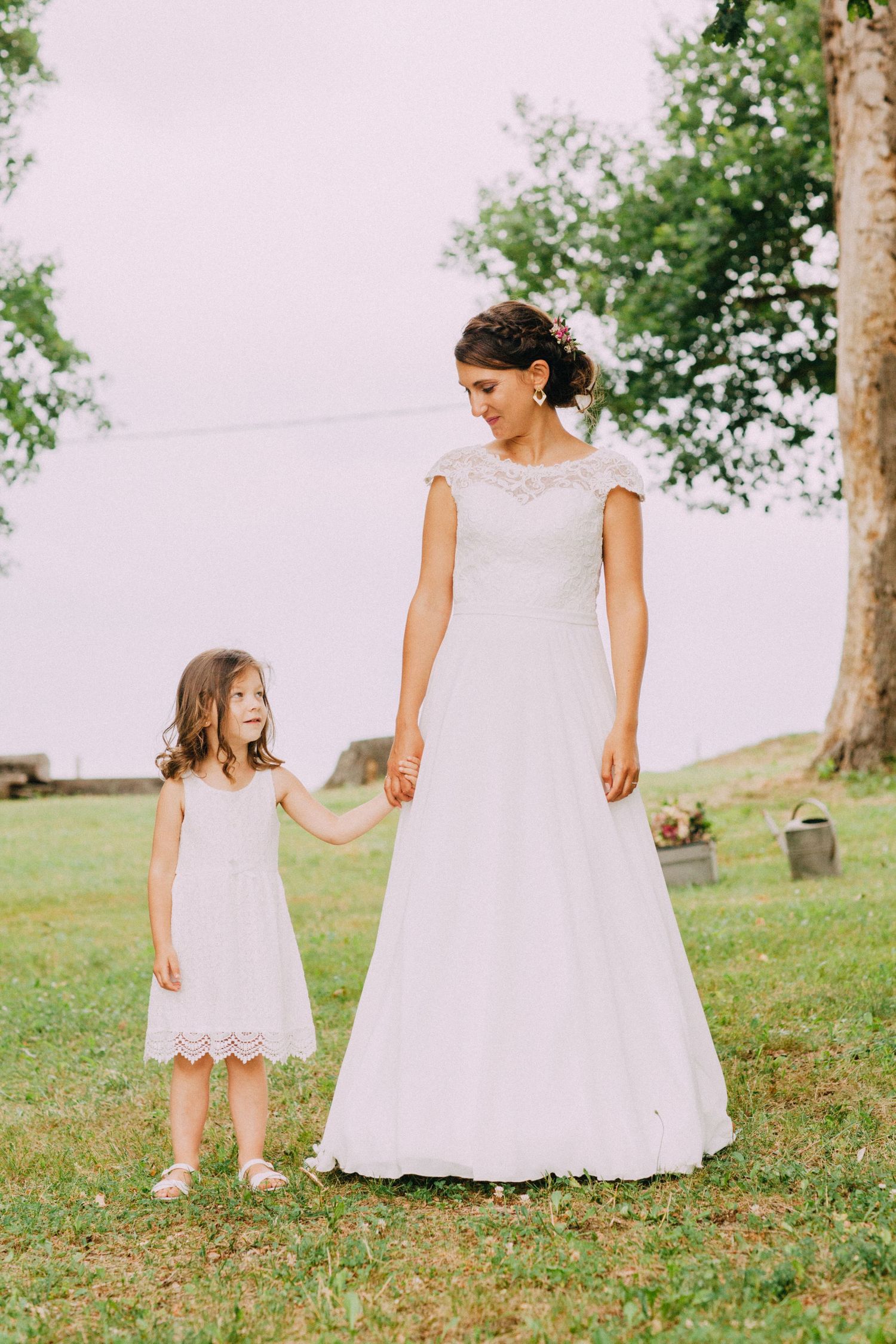 A bride in white lace gown stands with a young flower girl in matching dress on grass.