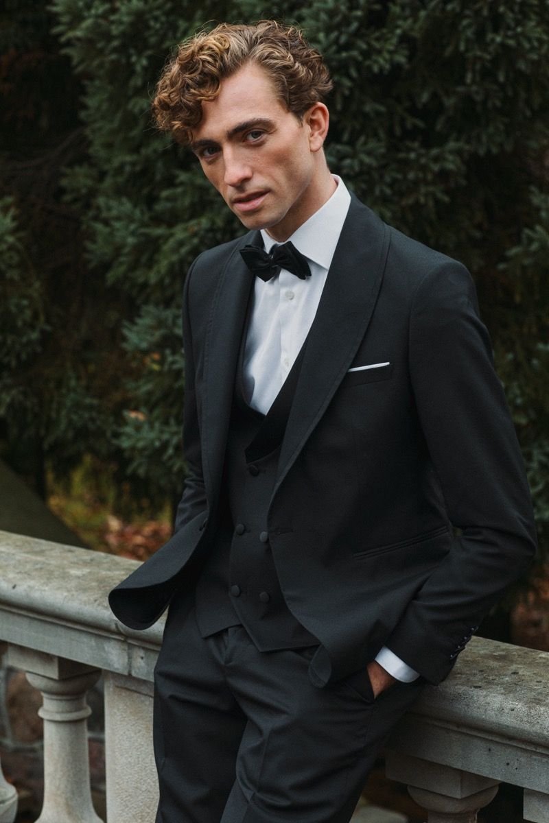 A groom in classic black tuxedo and bow tie poses against a garden backdrop.