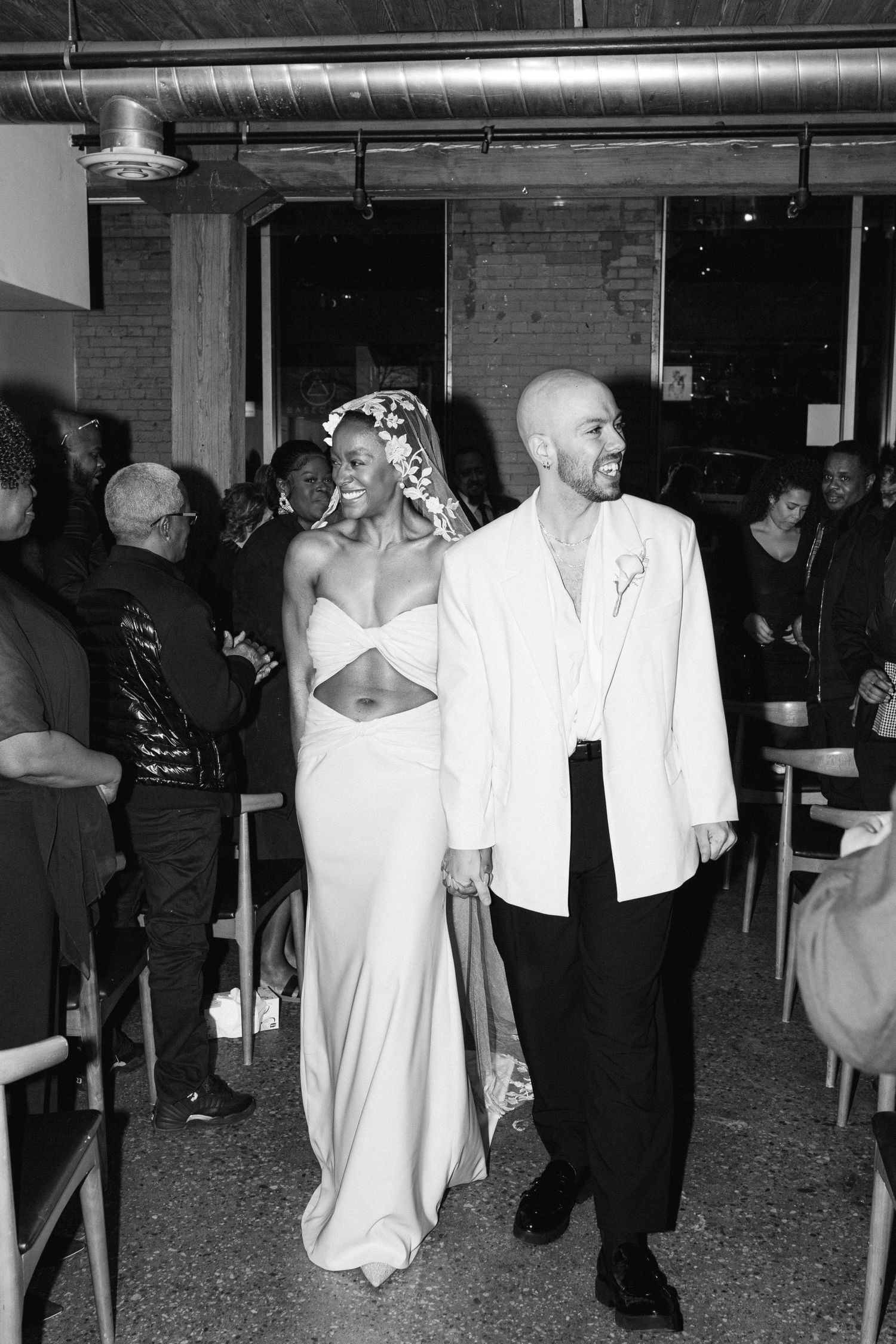 A wedding couple walks together through an industrial-style venue with exposed pipes and brick walls.