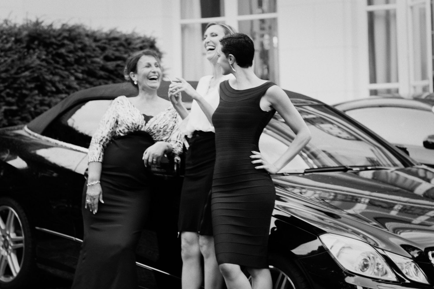 Two people in black dresses share a laugh next to a luxury car.
