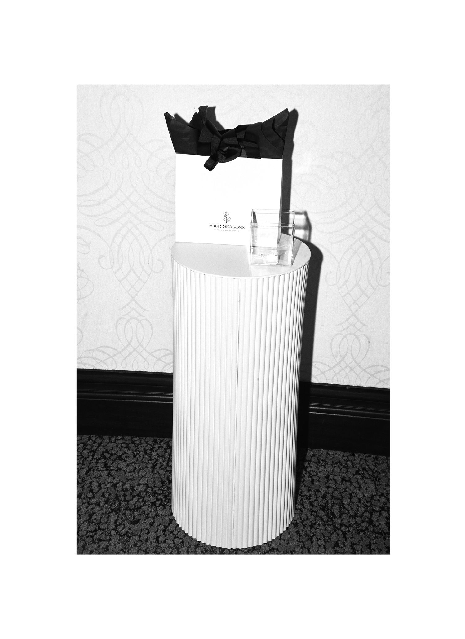Cylindrical white pedestal with black bow and shopping bag on top.