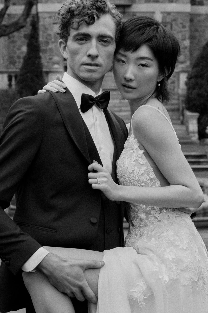 Elegant black and white wedding portrait of couple posing against stone building.