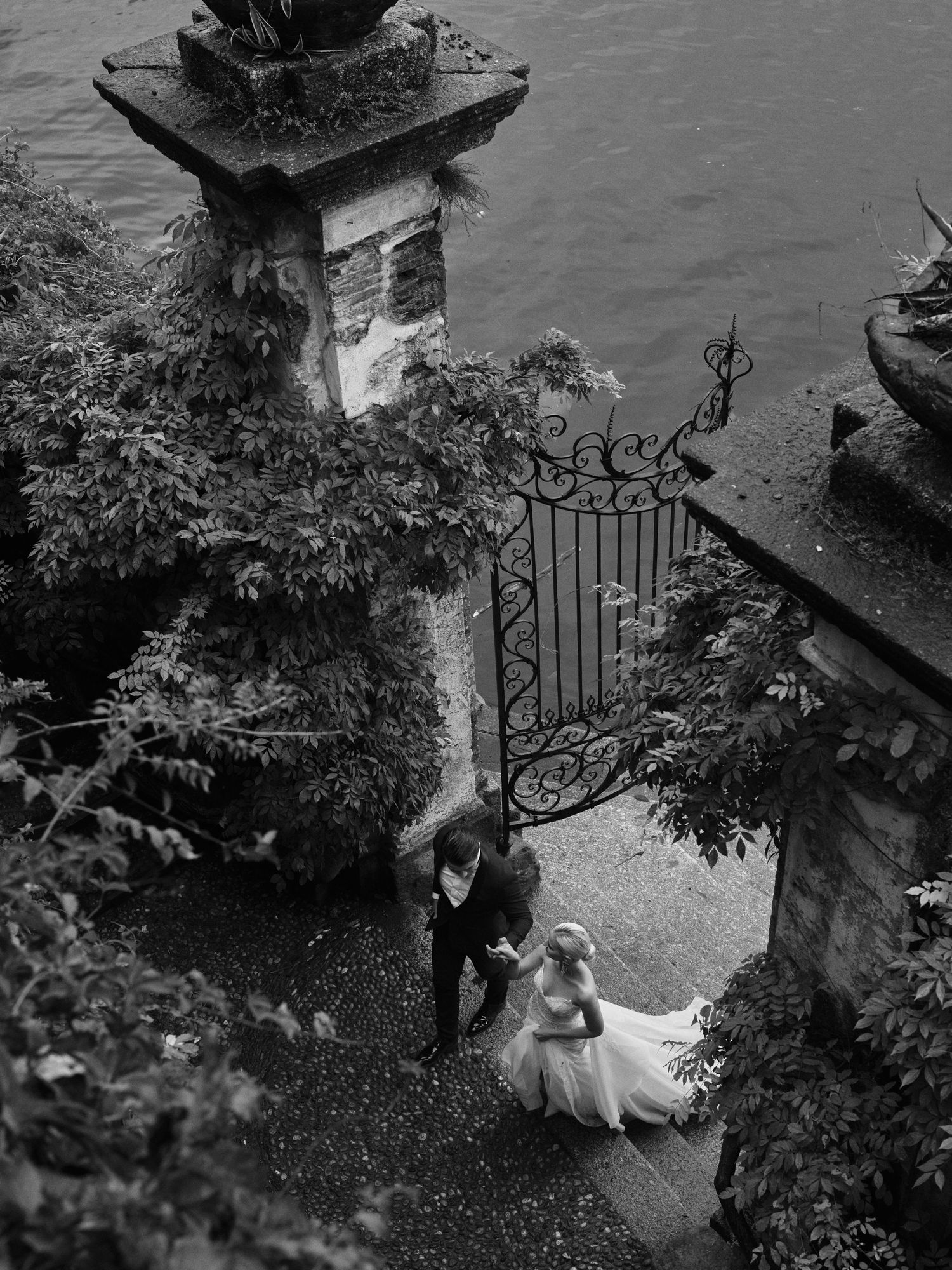 A black and white photograph of an ornate garden gate entrance with stone pillars and foliage by the water.