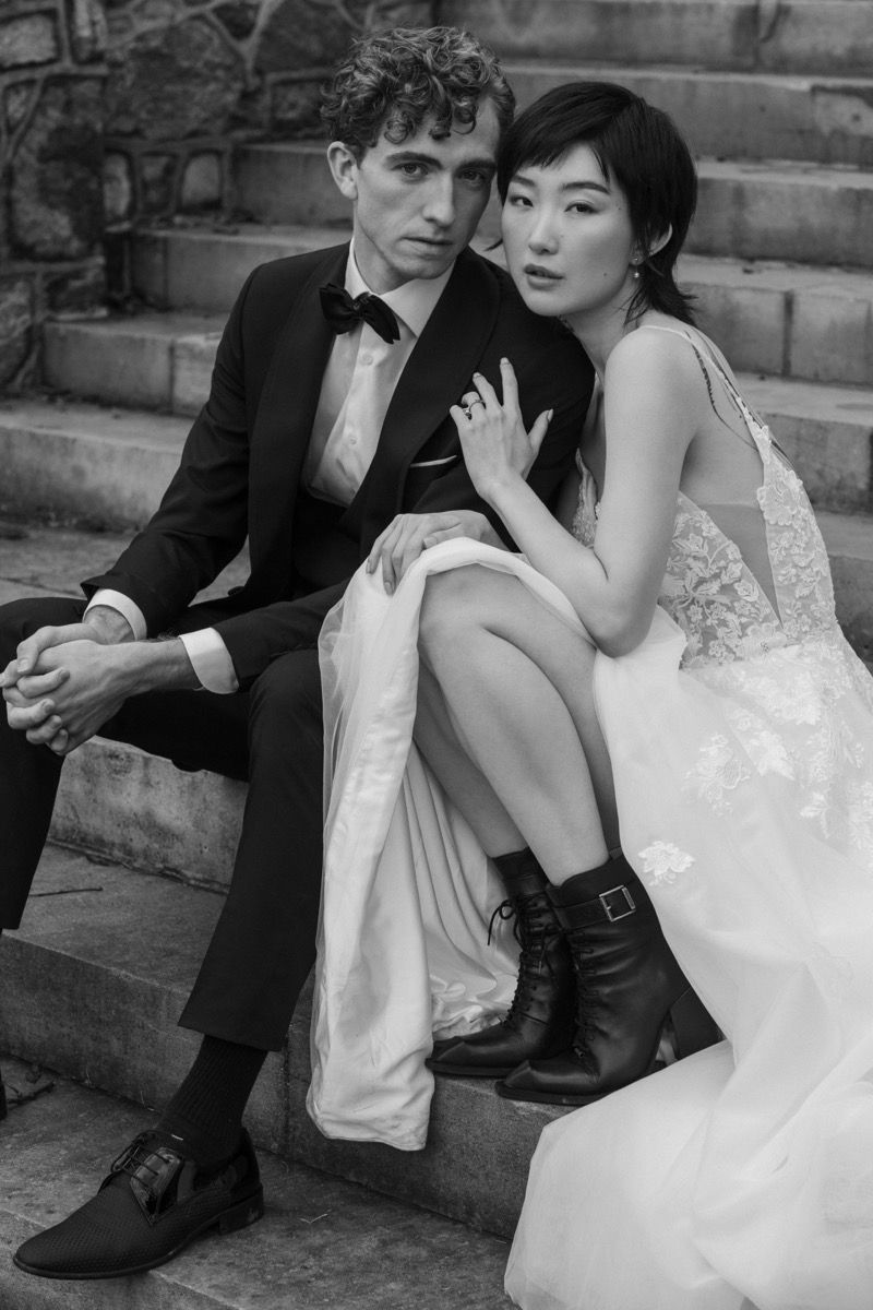 Stylish bride in white dress and boots sits with groom on stone steps in black and white portrait.