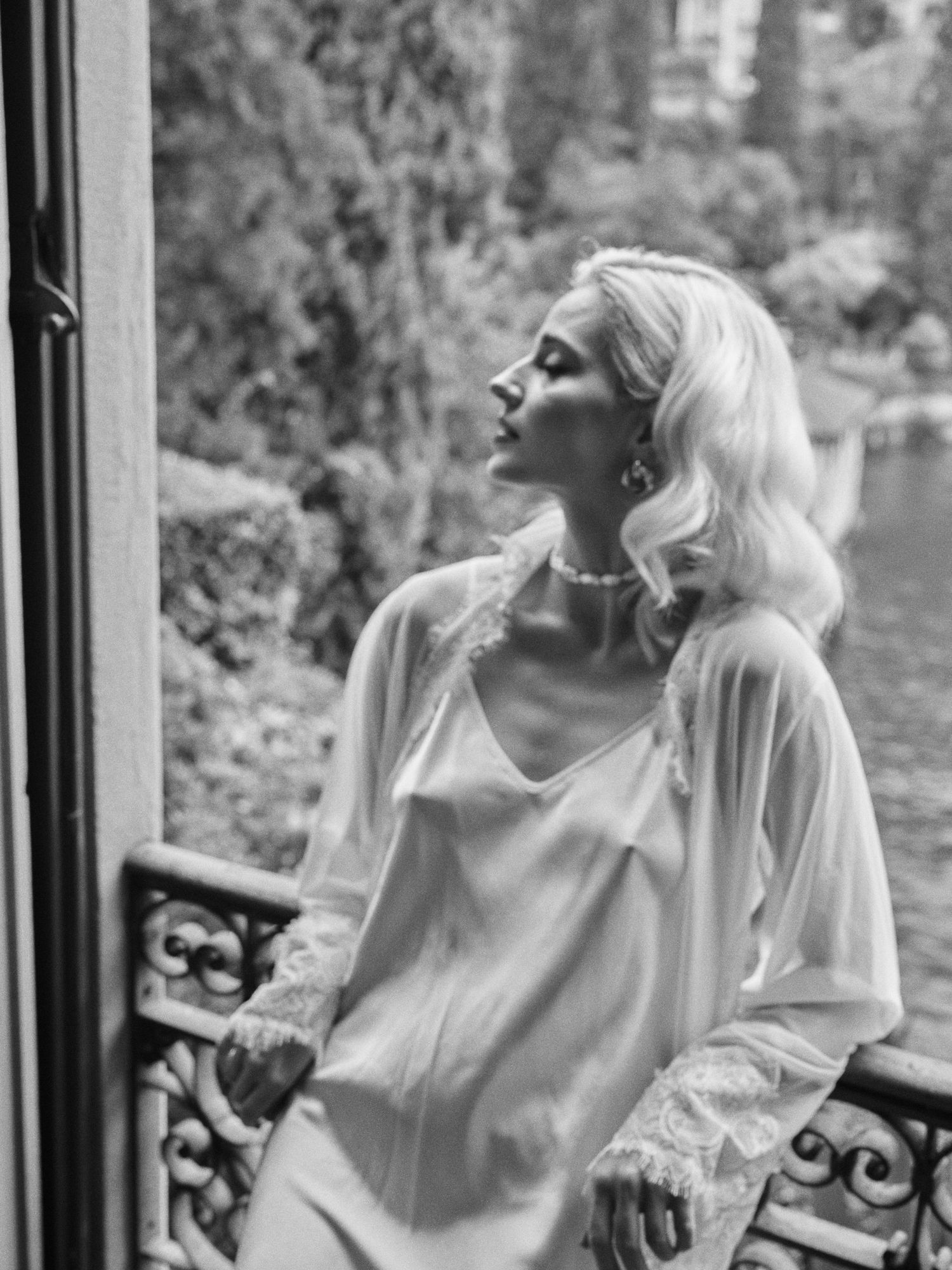 Black and white portrait of a woman in a white flowing garment looking up near an ornate balcony railing.