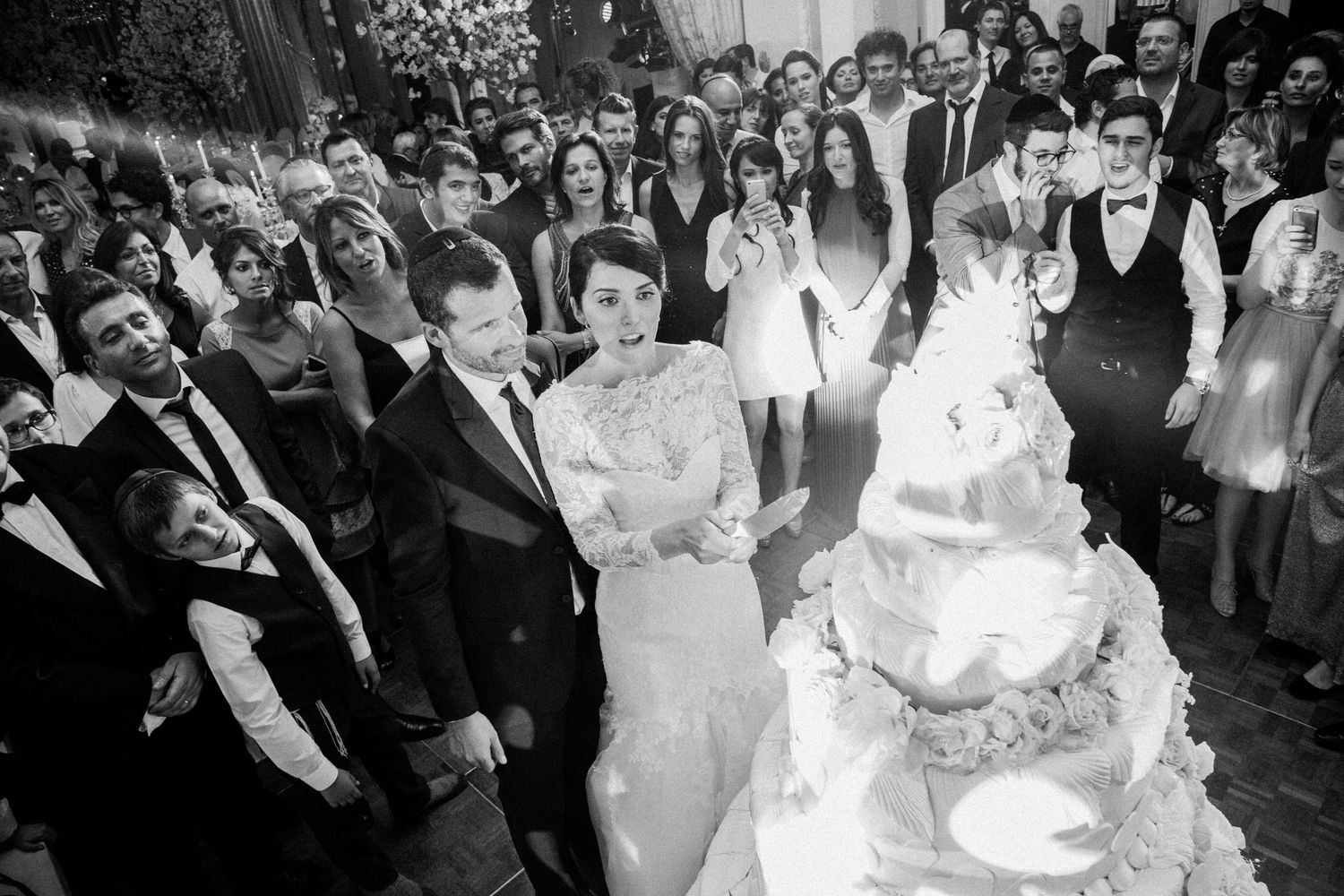 Wedding celebration scene showing a large crowd gathered around an elegant tiered white cake.