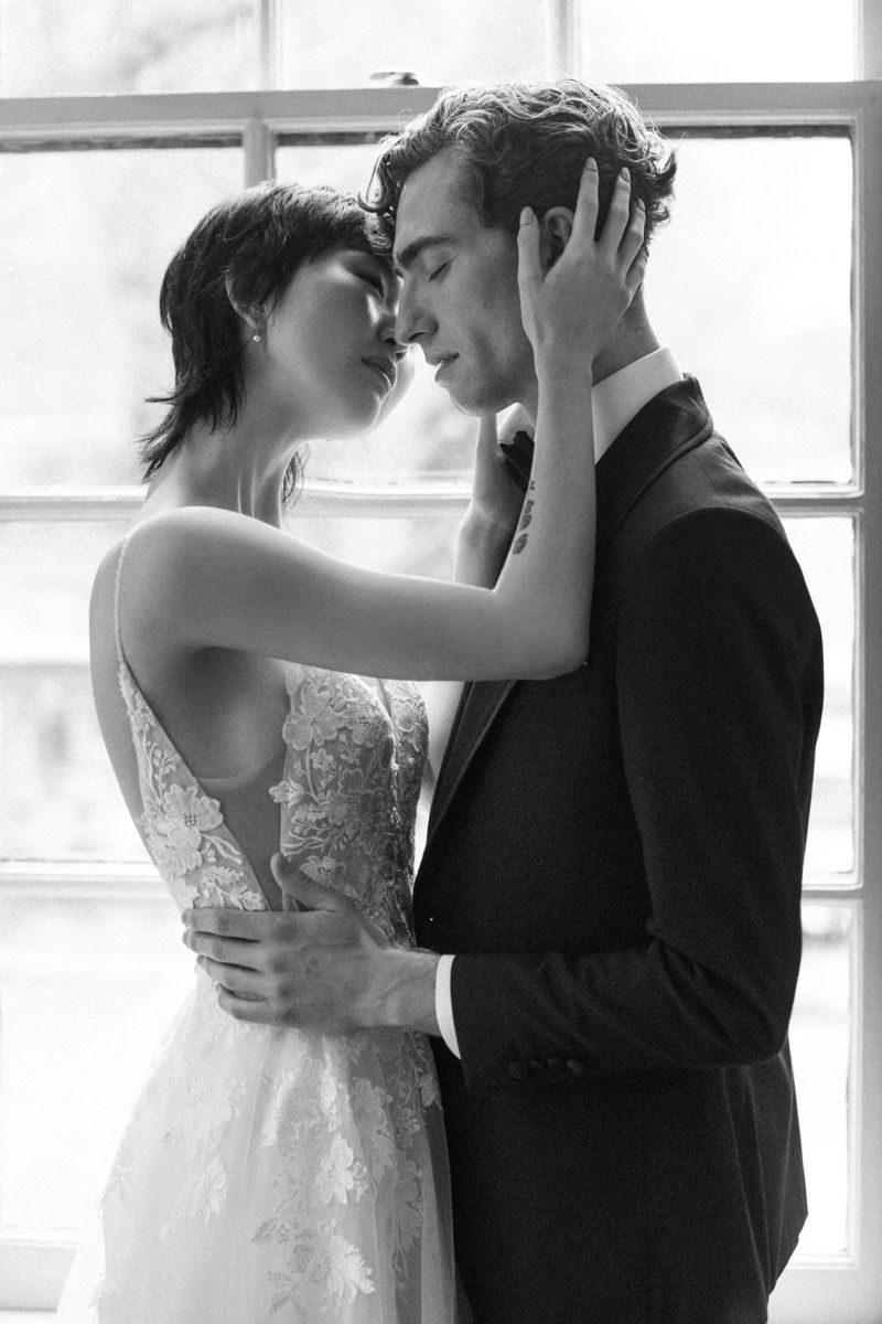 Intimate black and white portrait of a couple in wedding attire sharing a tender moment by a window.