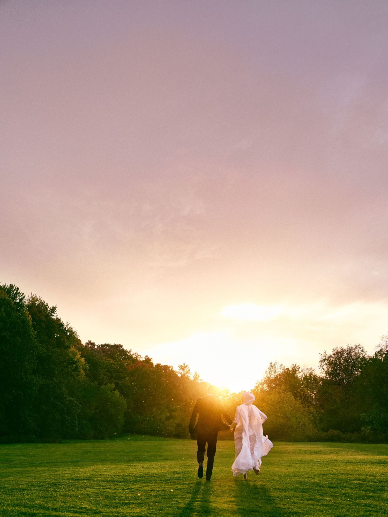 A couple walks hand in hand towards a golden sunset across a green field.