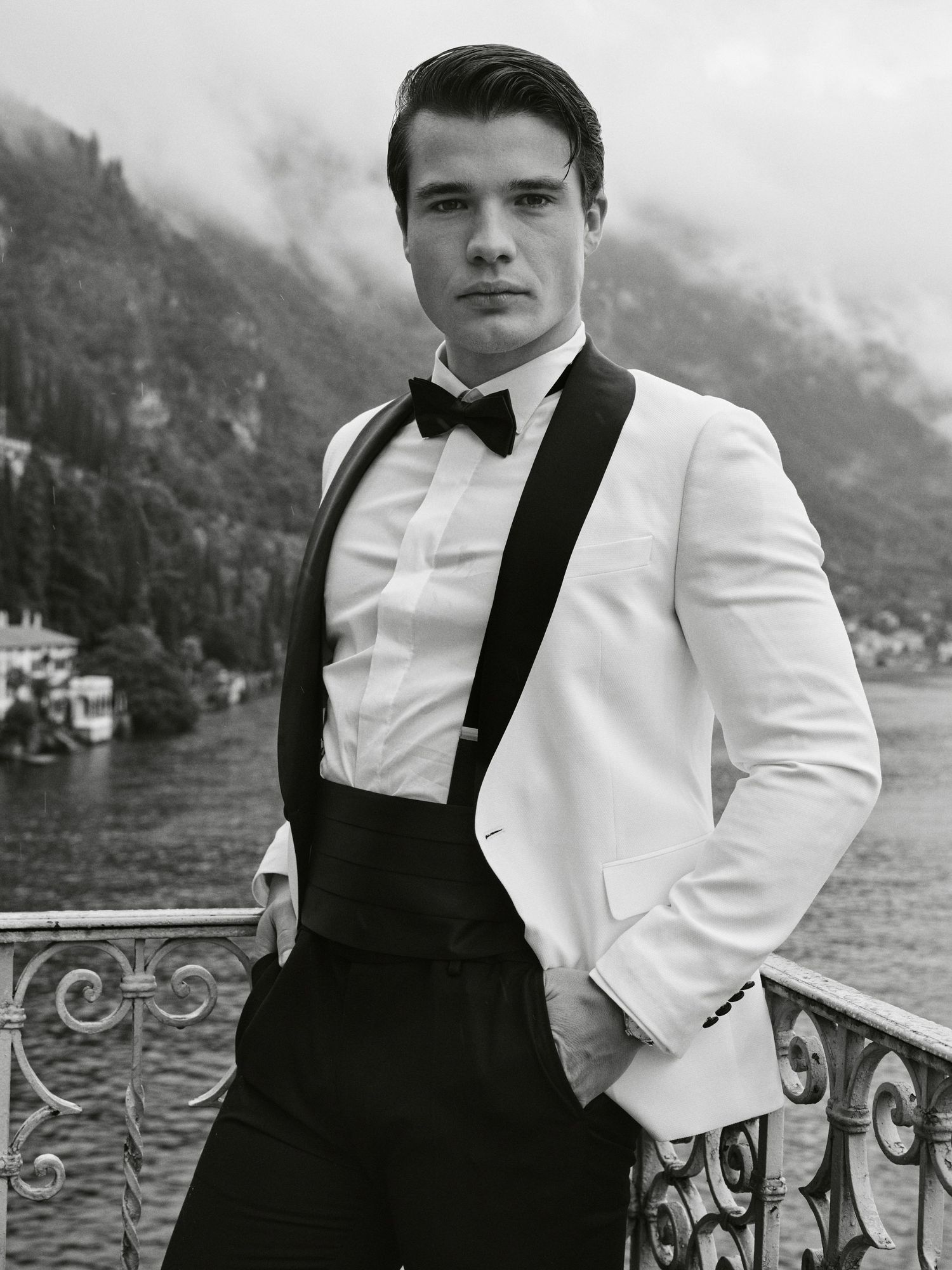 A formal portrait of someone in a white tuxedo jacket standing on a balcony overlooking a lake.