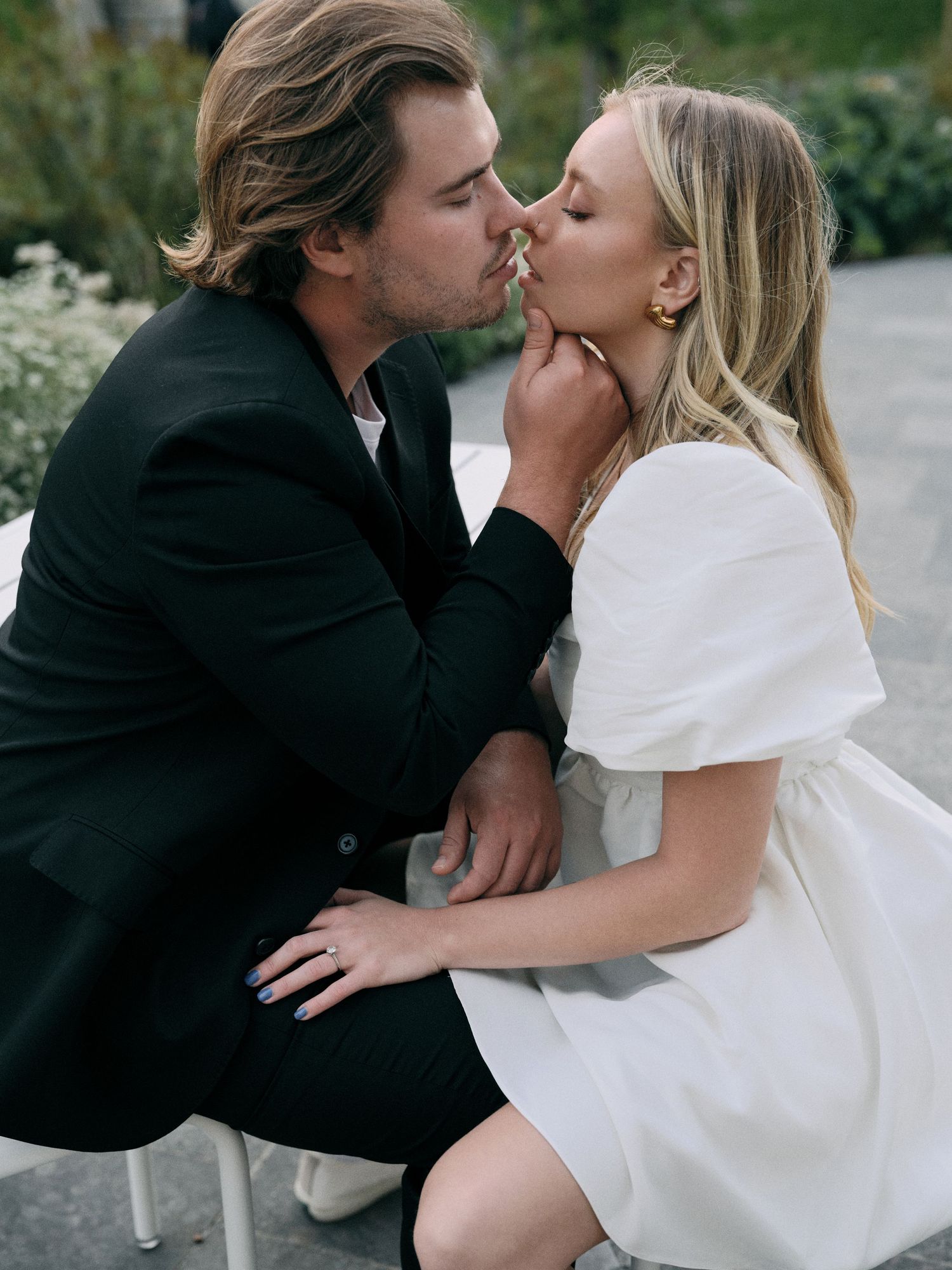 Romantic couple sharing an intimate kiss outdoors, with her in a white dress and him in a black jacket.