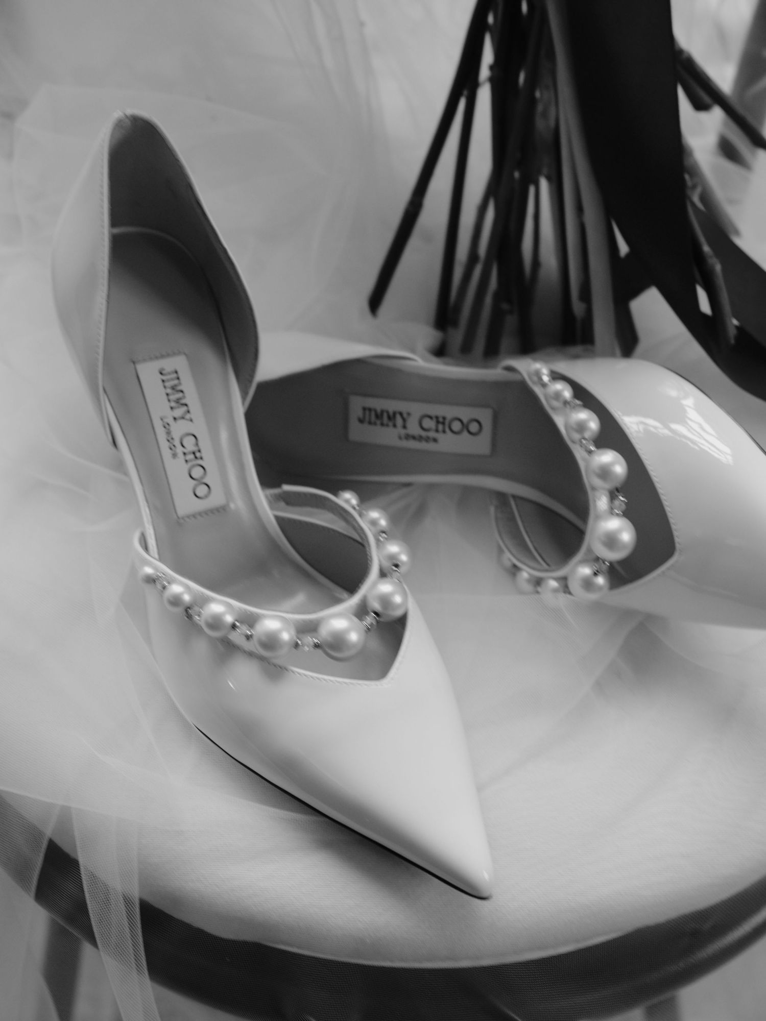 Designer Jimmy Choo bridal shoes in white satin with pearl embellishments displayed on tulle fabric in black and white.